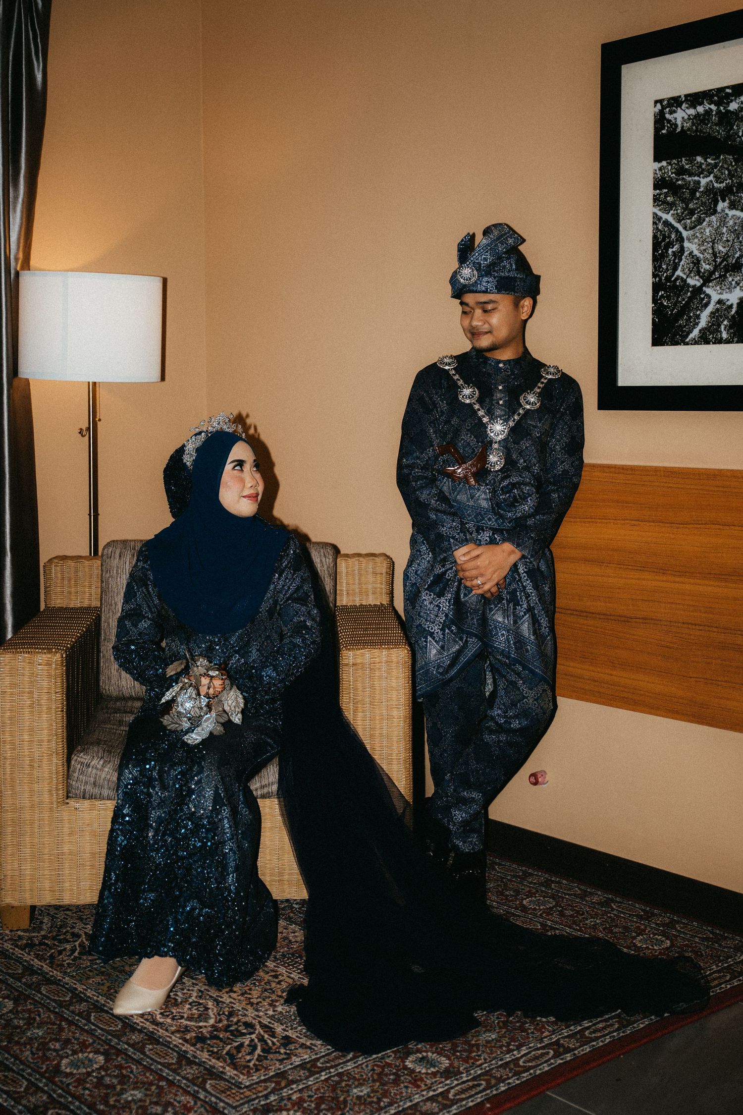 Two figures wearing dark kimono-style robes sit on a patterned carpet in a dimly lit room with beige walls.