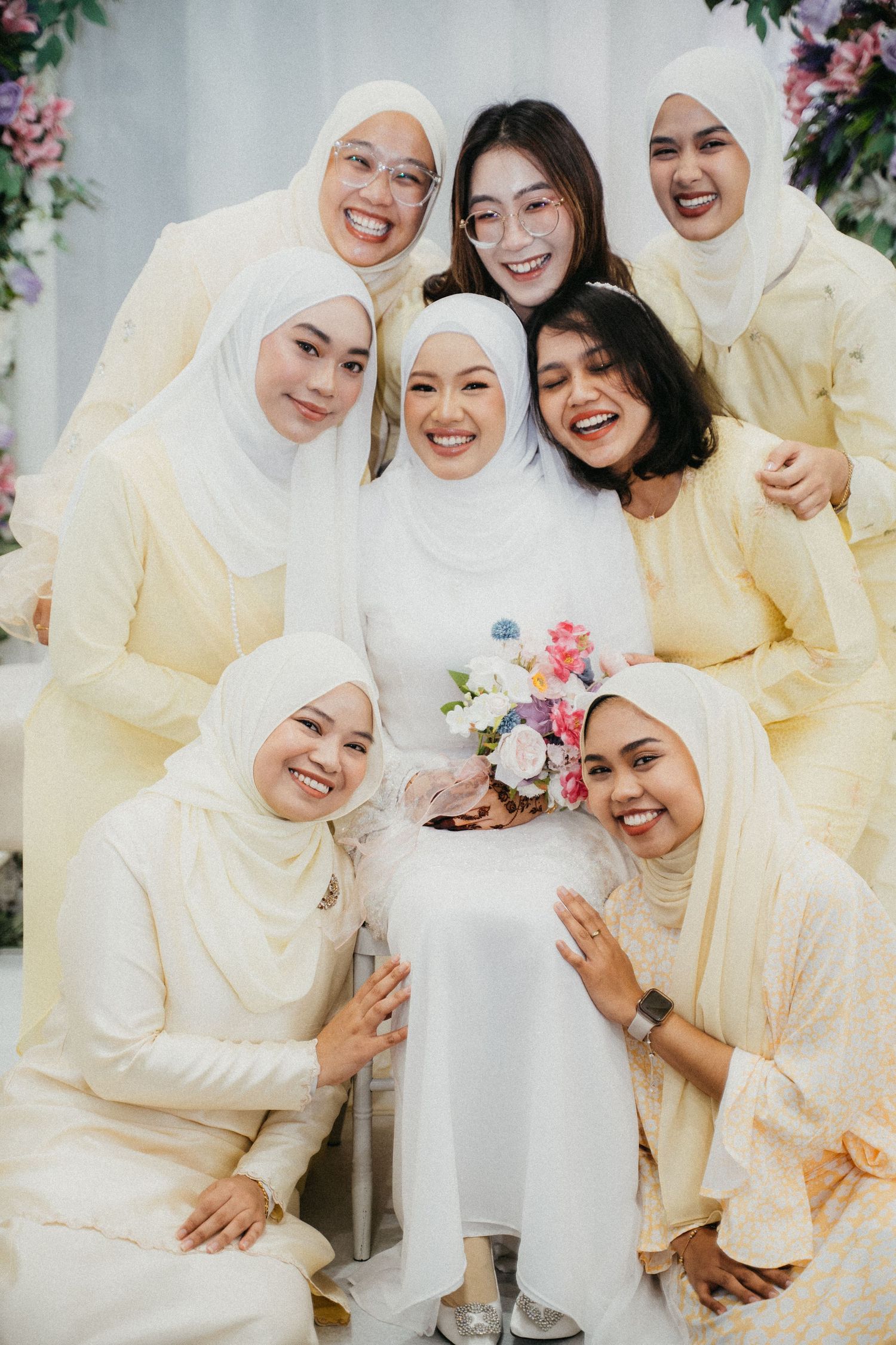Wedding party wearing traditional white and light yellow hijabs and dresses poses together with floral decorations.