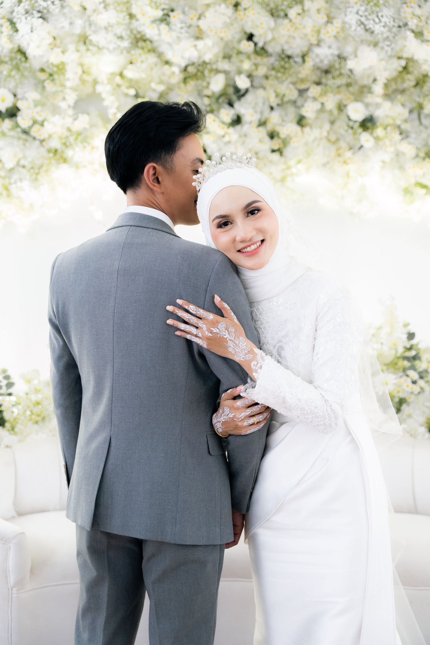 A couple in wedding attire embrace during a photoshoot with white floral decorations in the background.