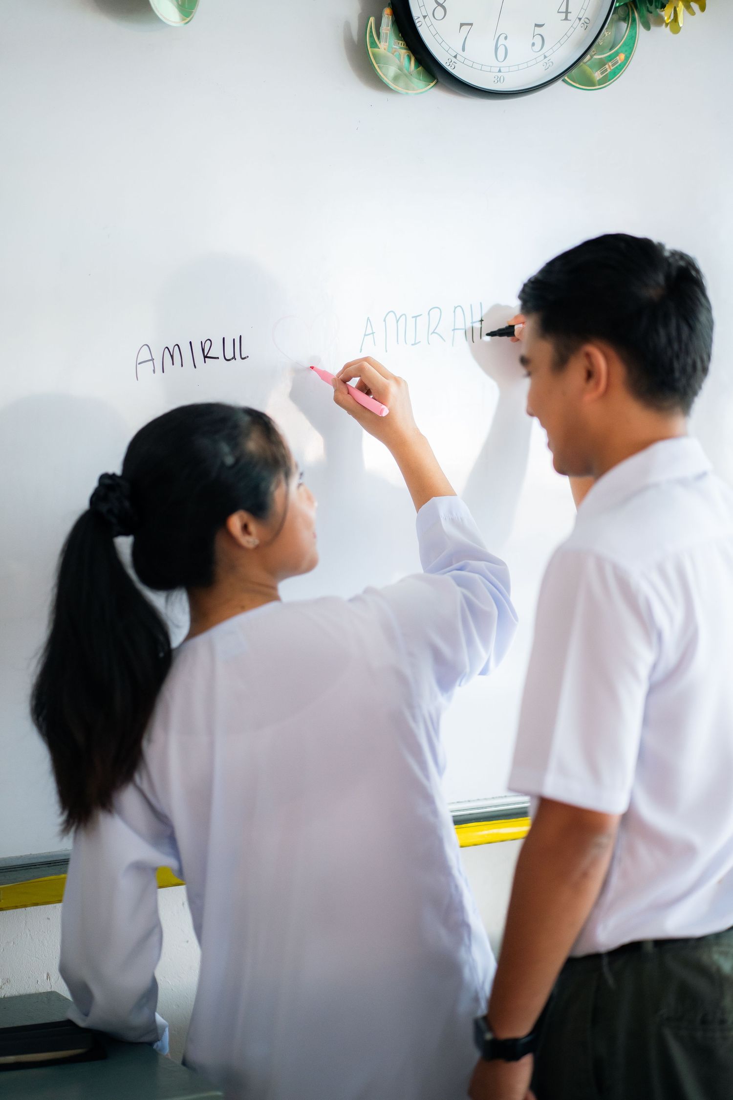 Students in white uniforms write on a whiteboard during a classroom learning session.