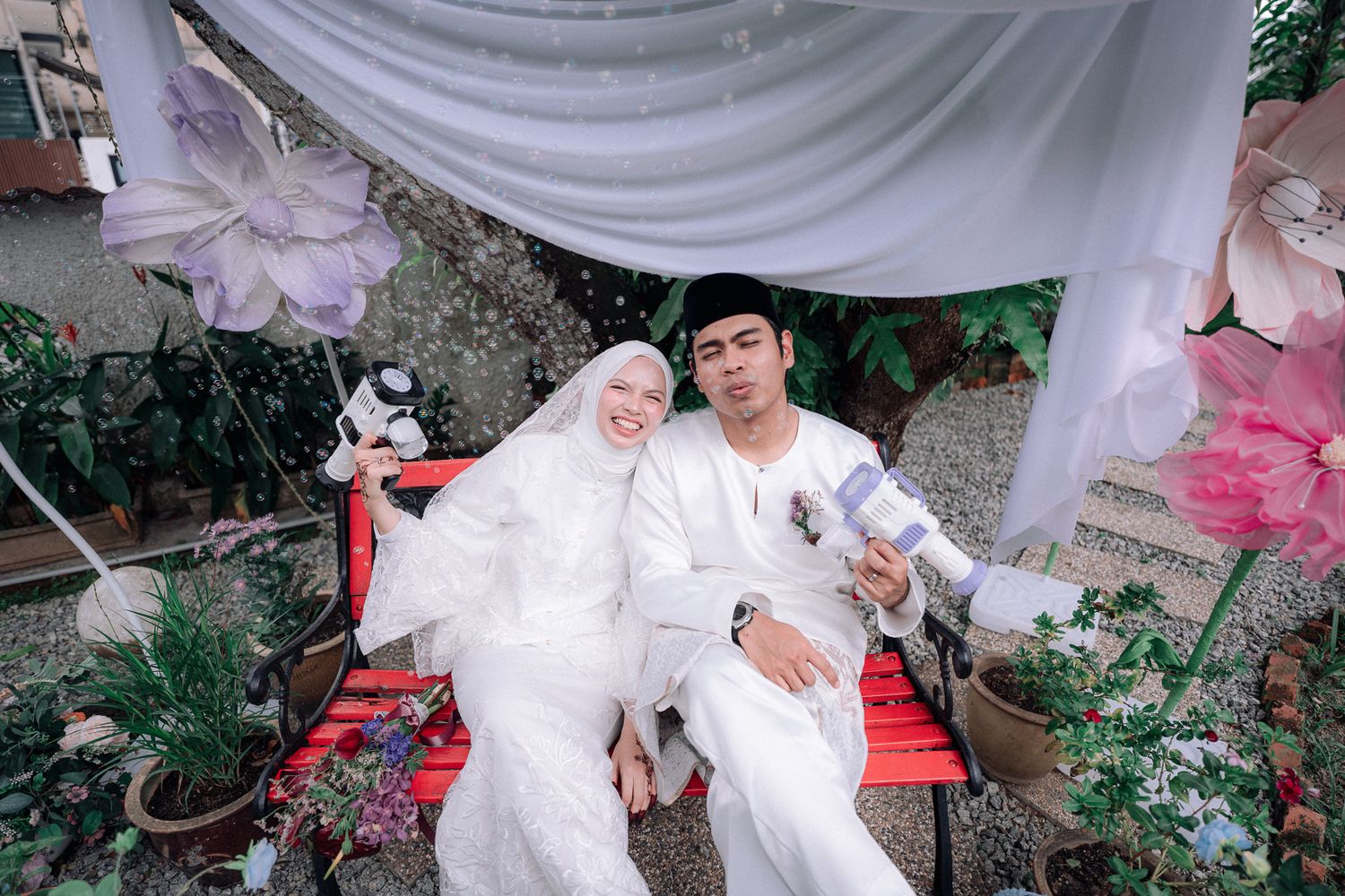 A couple in white traditional Muslim wedding attire poses on a red bench surrounded by lush greenery and floral decorations.