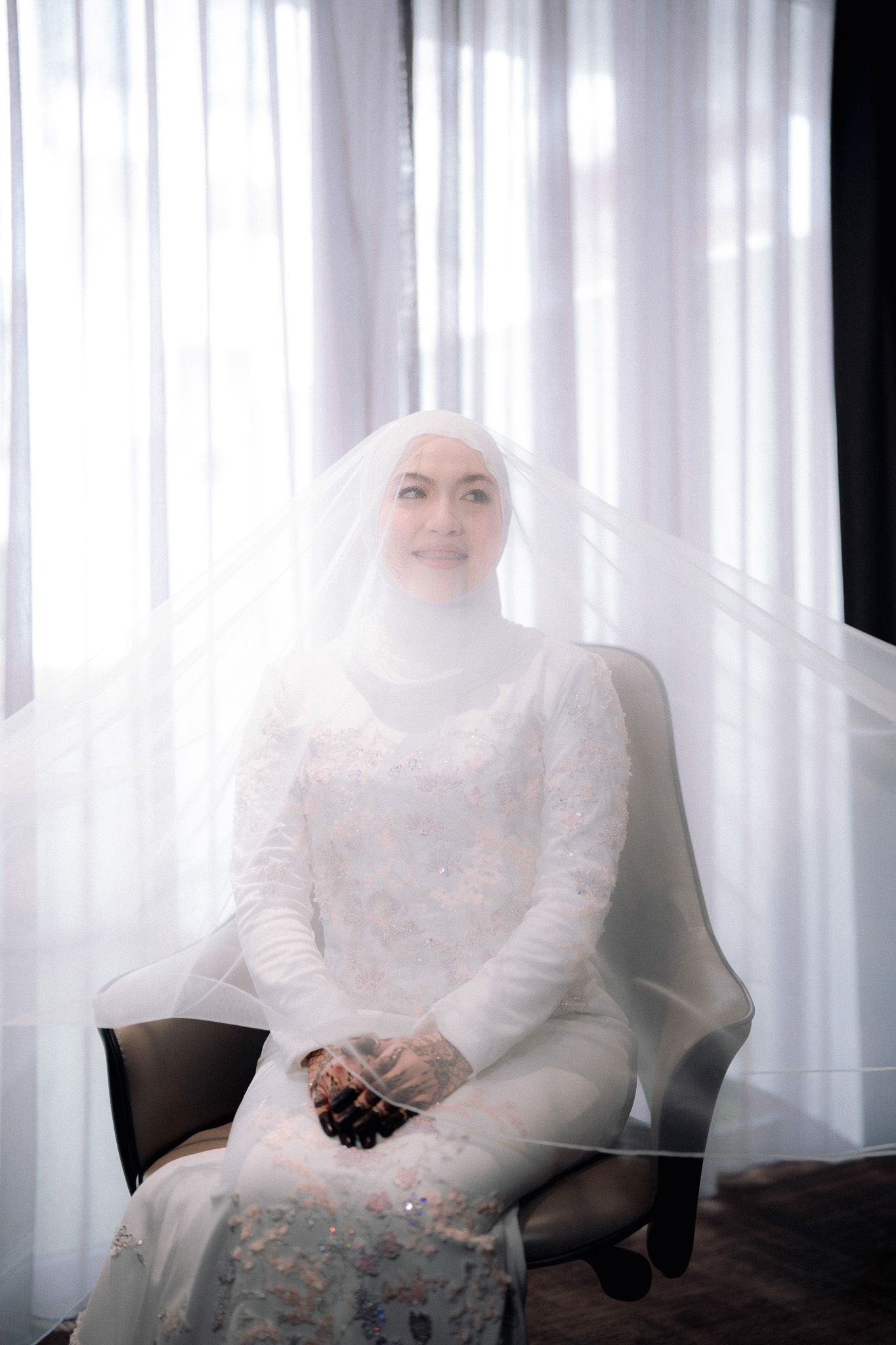 Muslim bride in white hijab and lace wedding dress seated by bright window with veil.