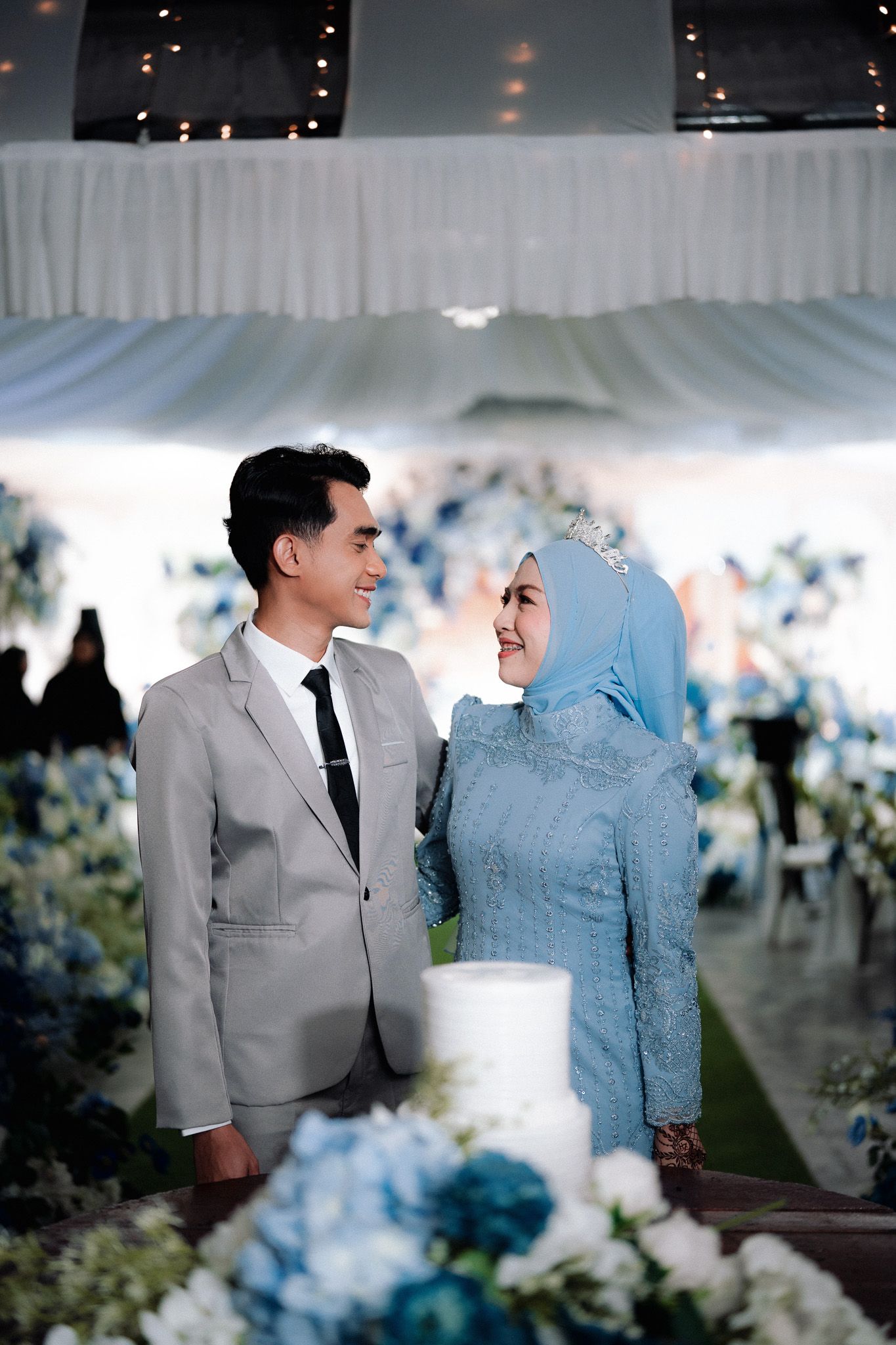 Muslim couple in blue and gray wedding attire smiling at each other beside a white cake and blue floral arrangements.