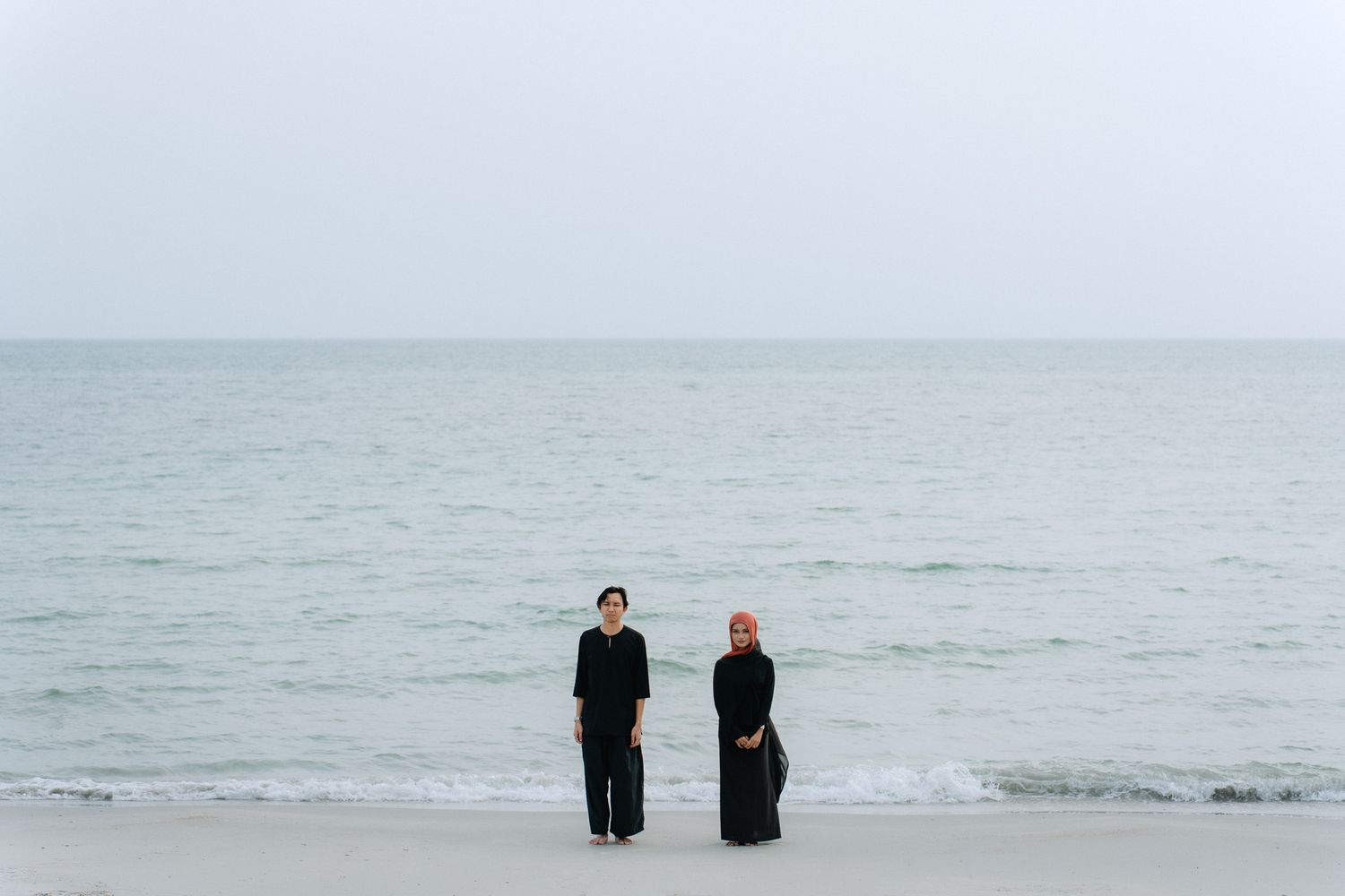 Two women in black outfits stand on a serene beach with calm ocean waves and overcast sky behind them.
