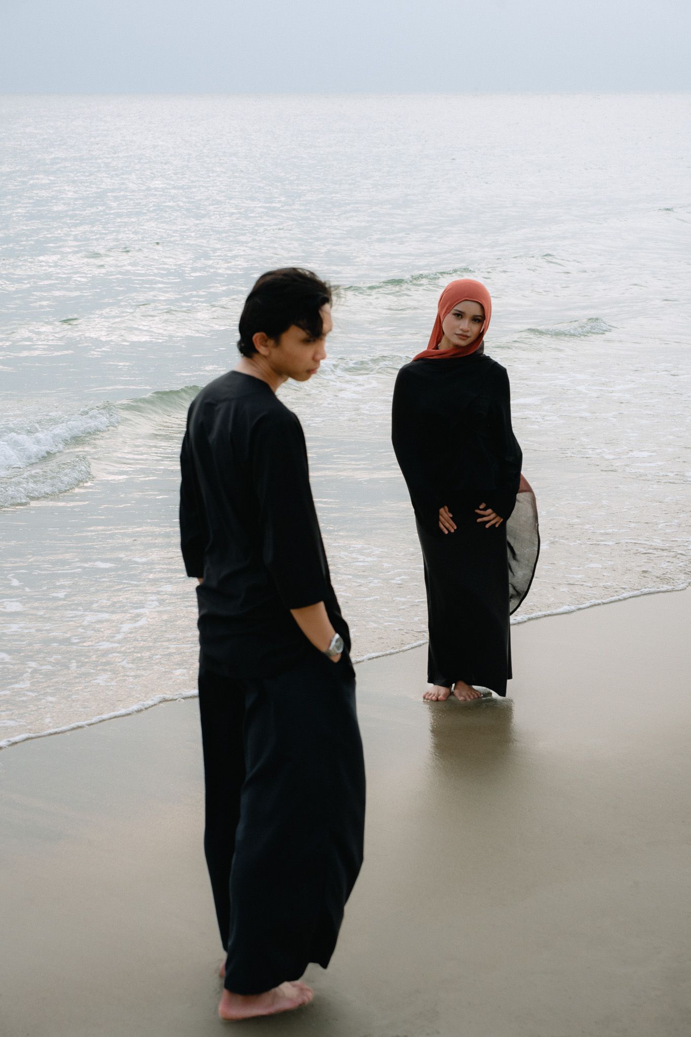 Man and woman in black outfits on sandy beach, woman in red hijab holding a fan by the ocean shore.