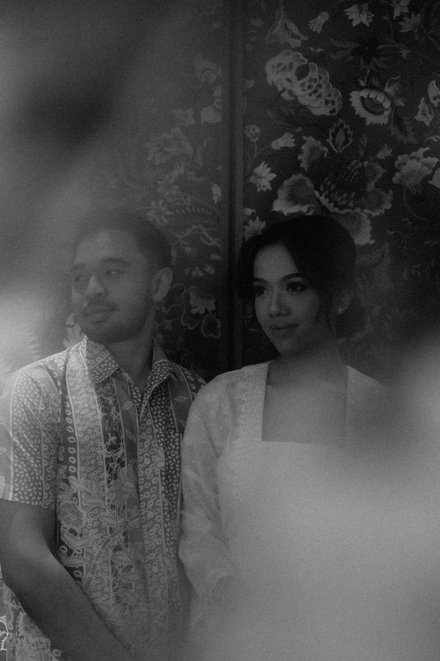 Black and white artistic portrait of two figures against a floral patterned wallpaper background.
