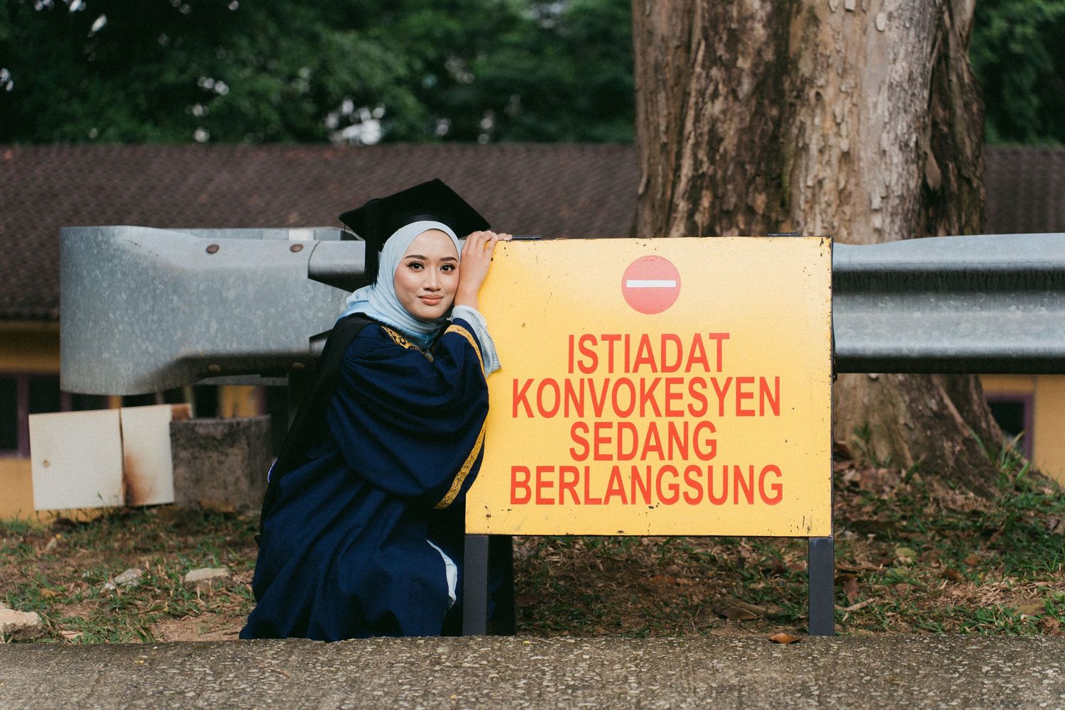 Person in black clothing sits next to yellow warning sign with Indonesian text on roadside near tree.
