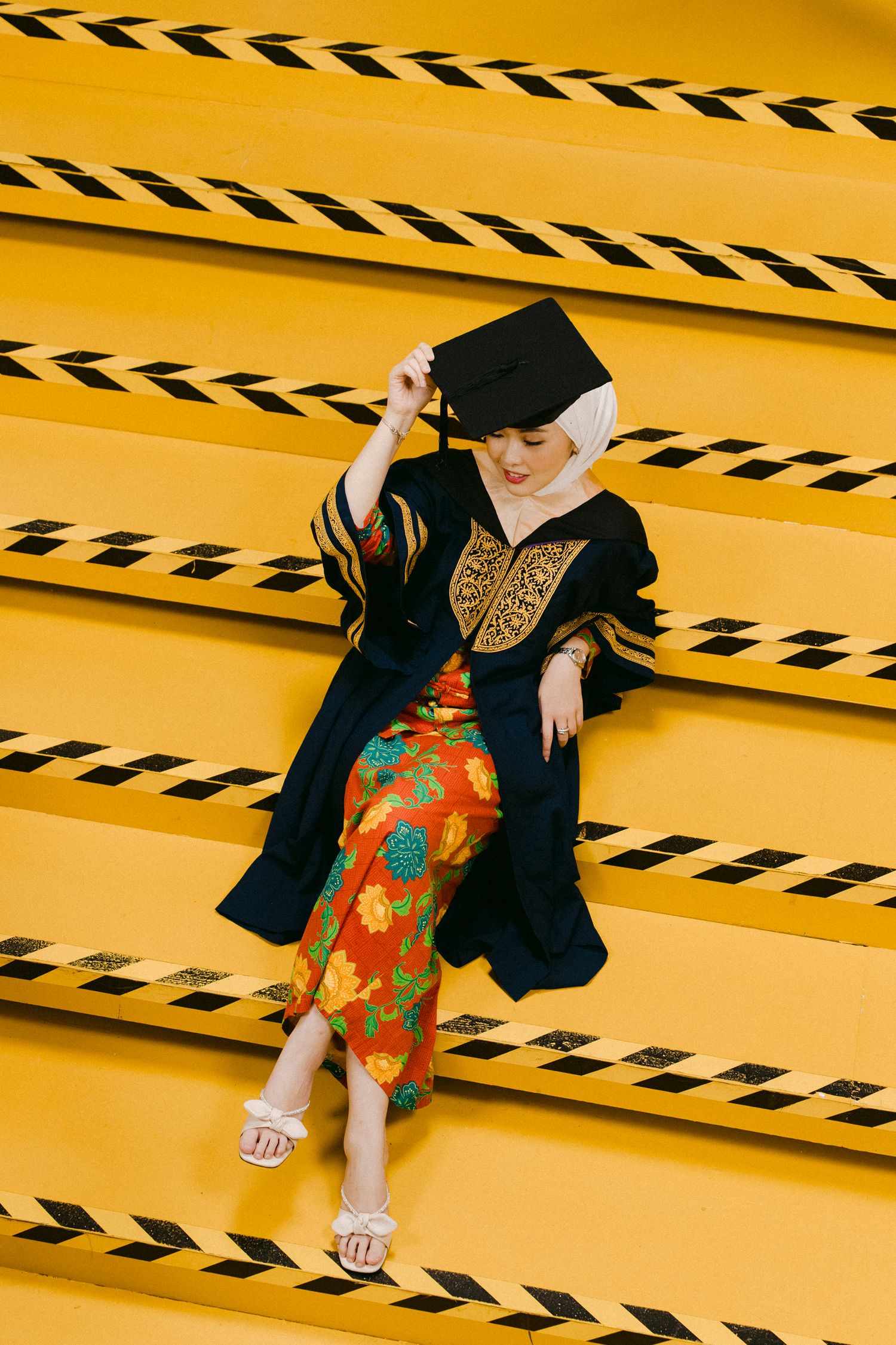 Graduate in traditional robe and floral dress posing on yellow steps with black stripe pattern.