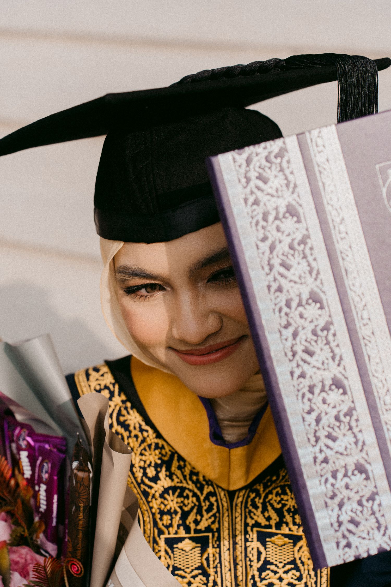 Graduate wearing black cap and yellow patterned graduation gown smiles while holding scroll at graduation ceremony.