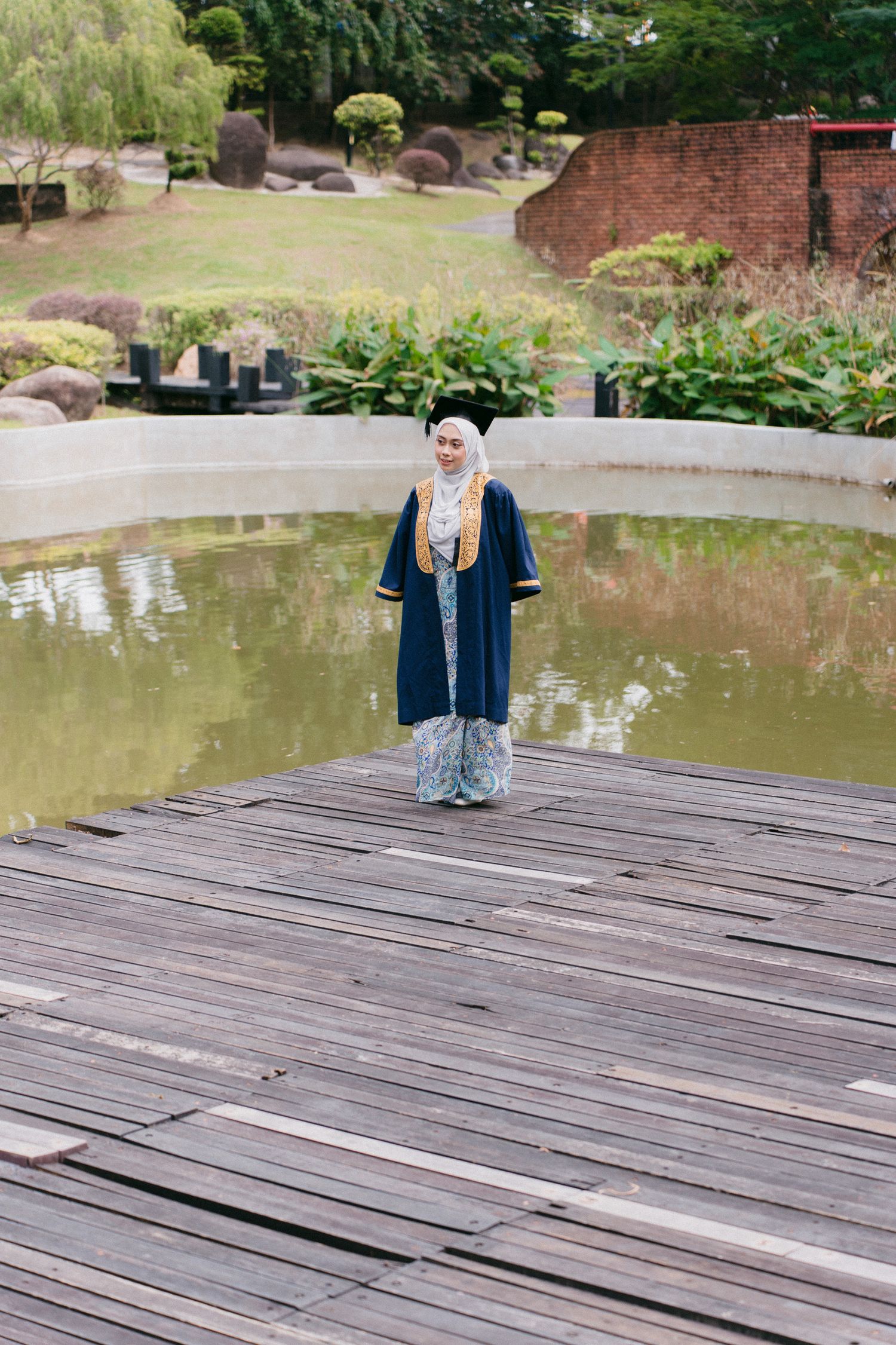 A person in a hijab and navy cardigan sits on a wooden deck overlooking a serene pond in a garden setting.