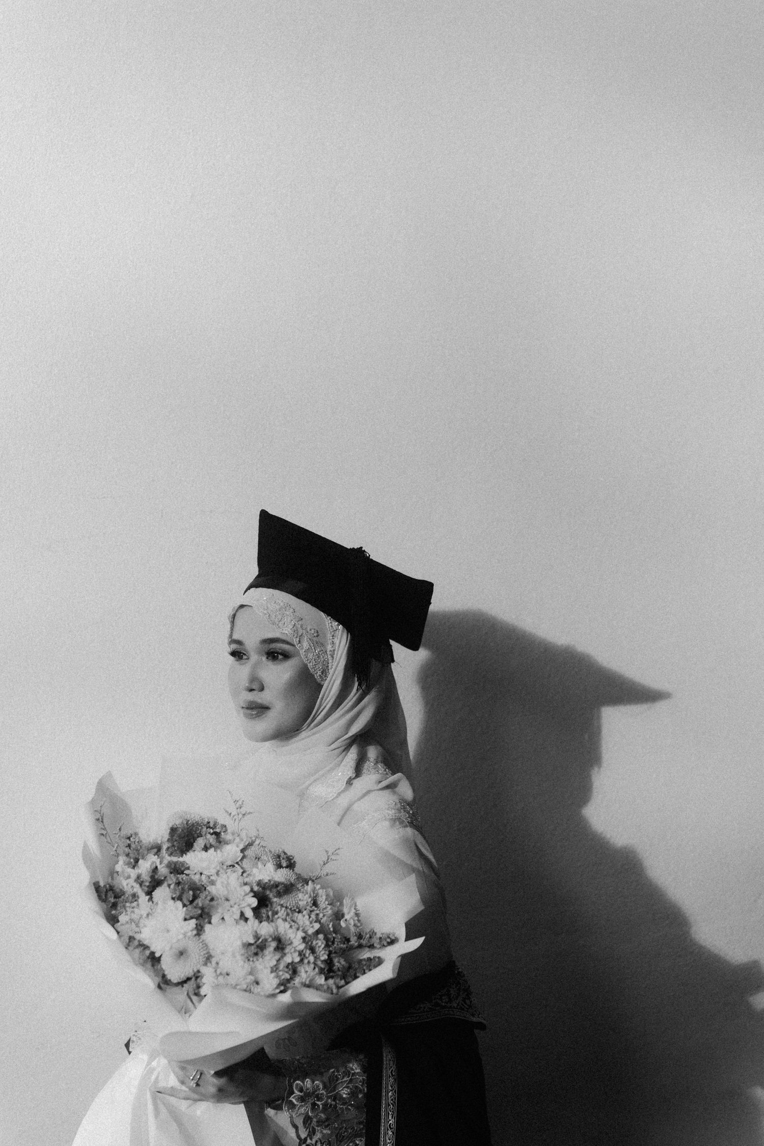 Black and white portrait of a graduate in cap and gown holding a floral bouquet against a light background.