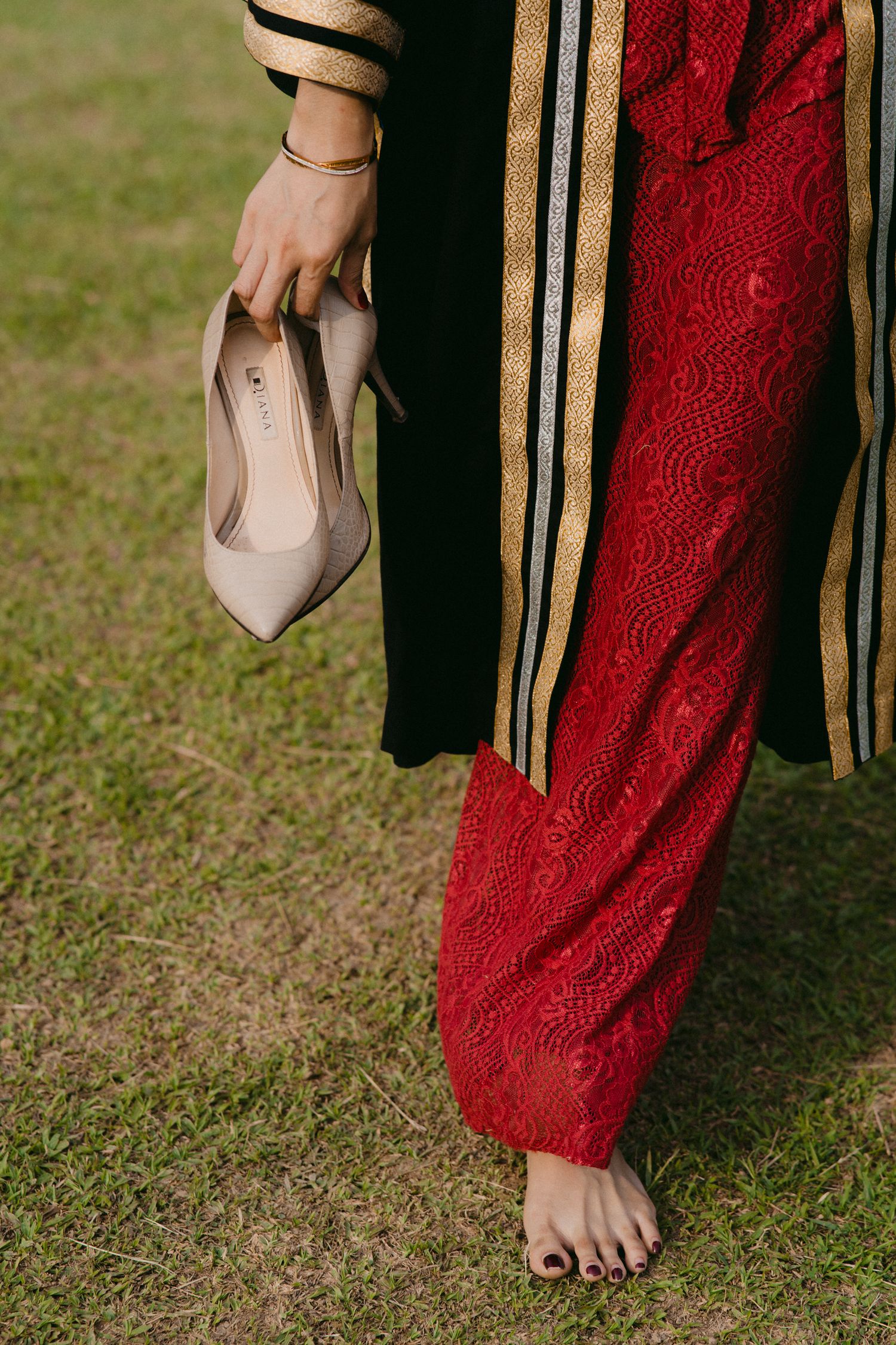 A hand holds a nude colored flat shoe while wearing red traditional pants with gold stripes on a grassy background.