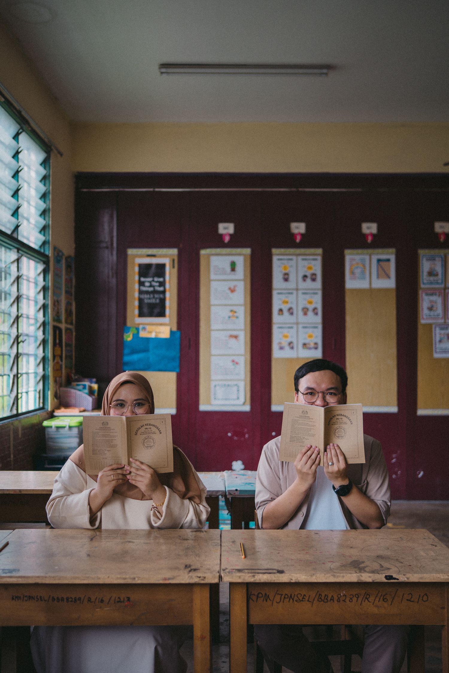 Two students hiding their faces behind books at wooden school desks.