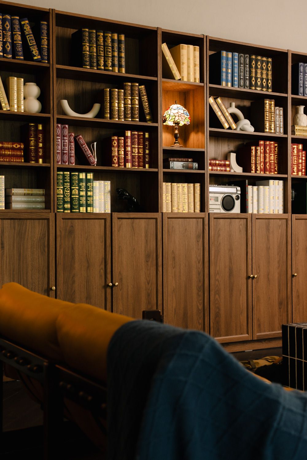 Elegant wooden bookshelf filled with classic books, a vintage radio, and a Tiffany lamp in a cozy library setting.