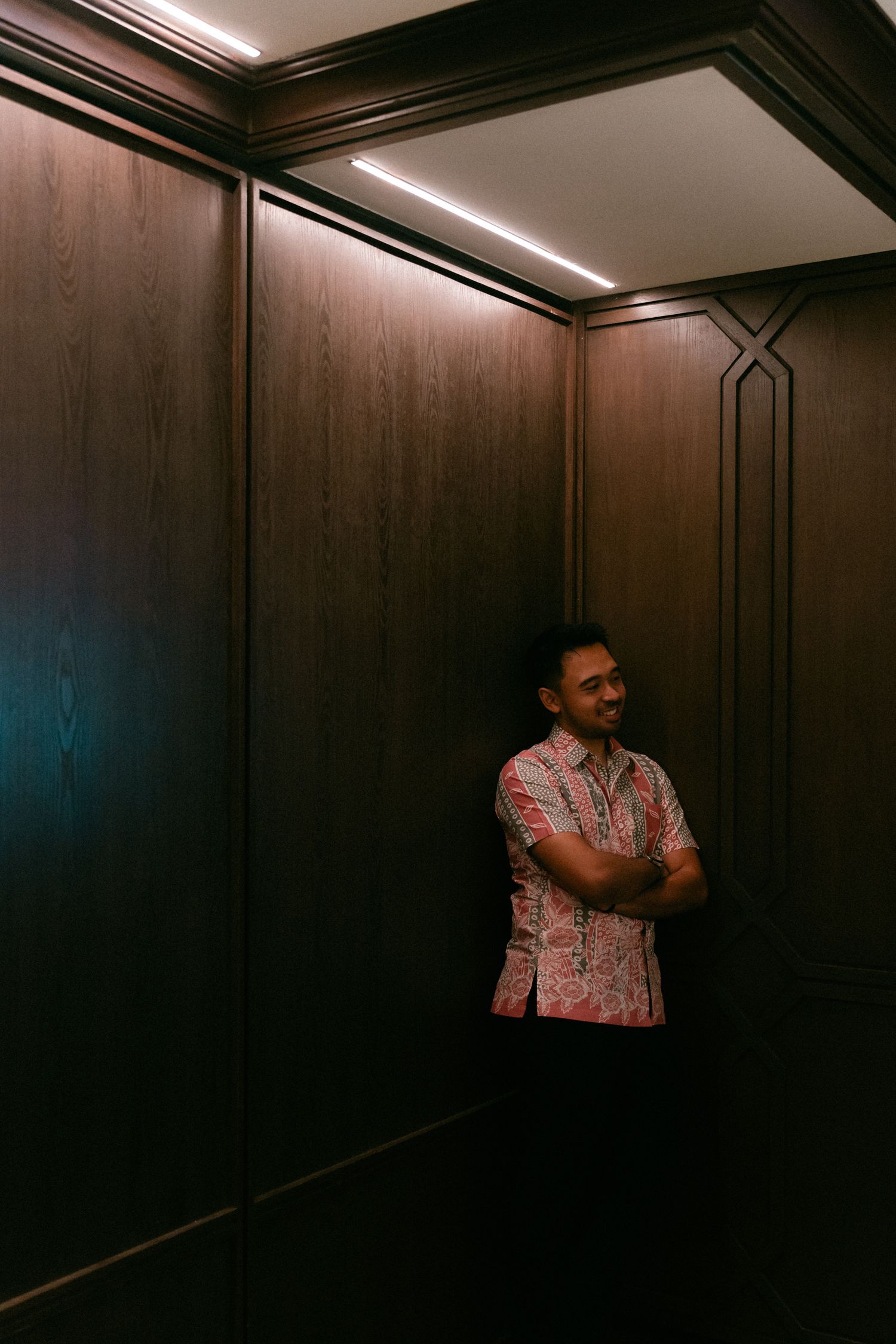 Person wearing a pink patterned shirt smiles while standing in the corner of a wood-paneled elevator.