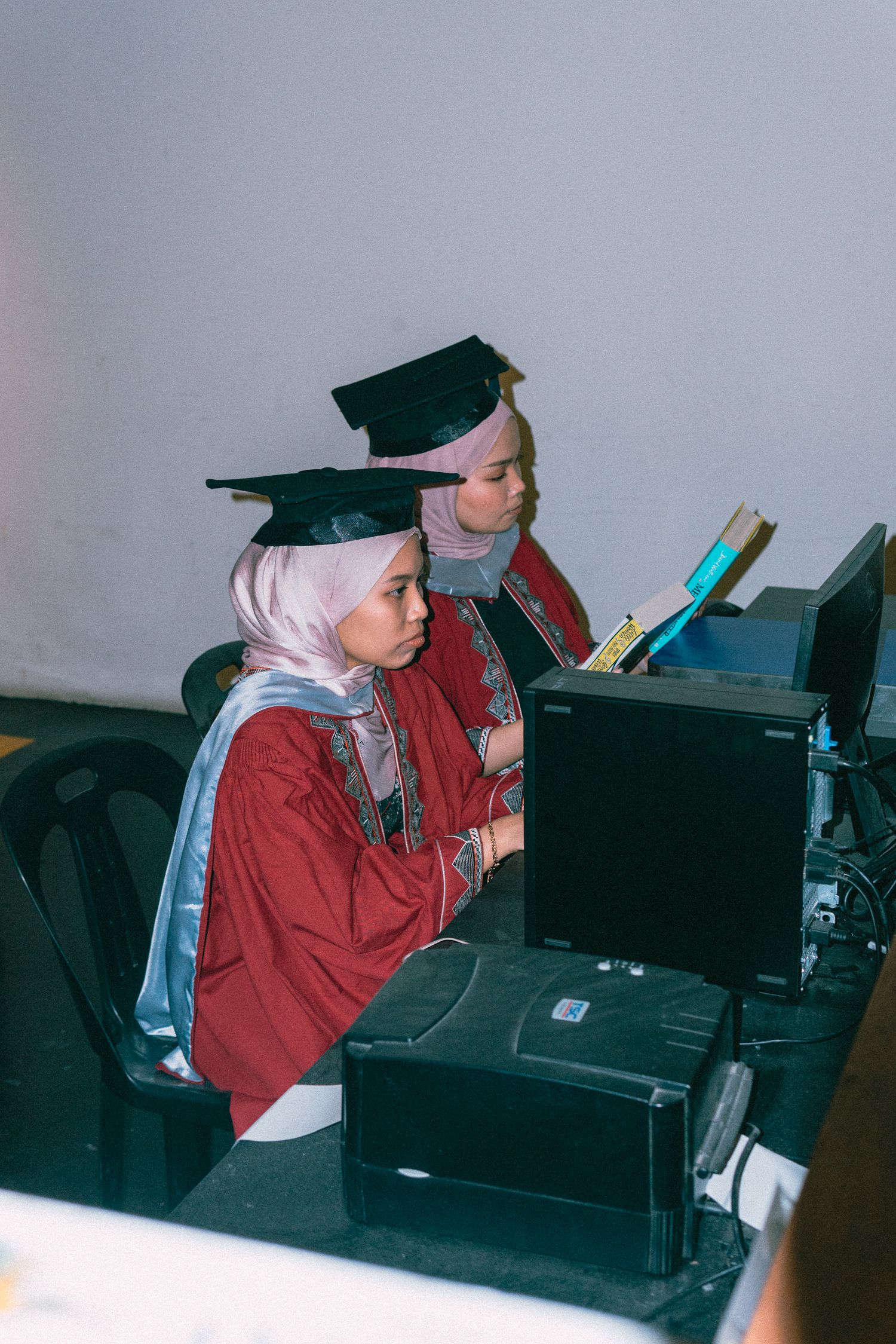 Two graduates in red robes work at computer stations during a graduation ceremony.