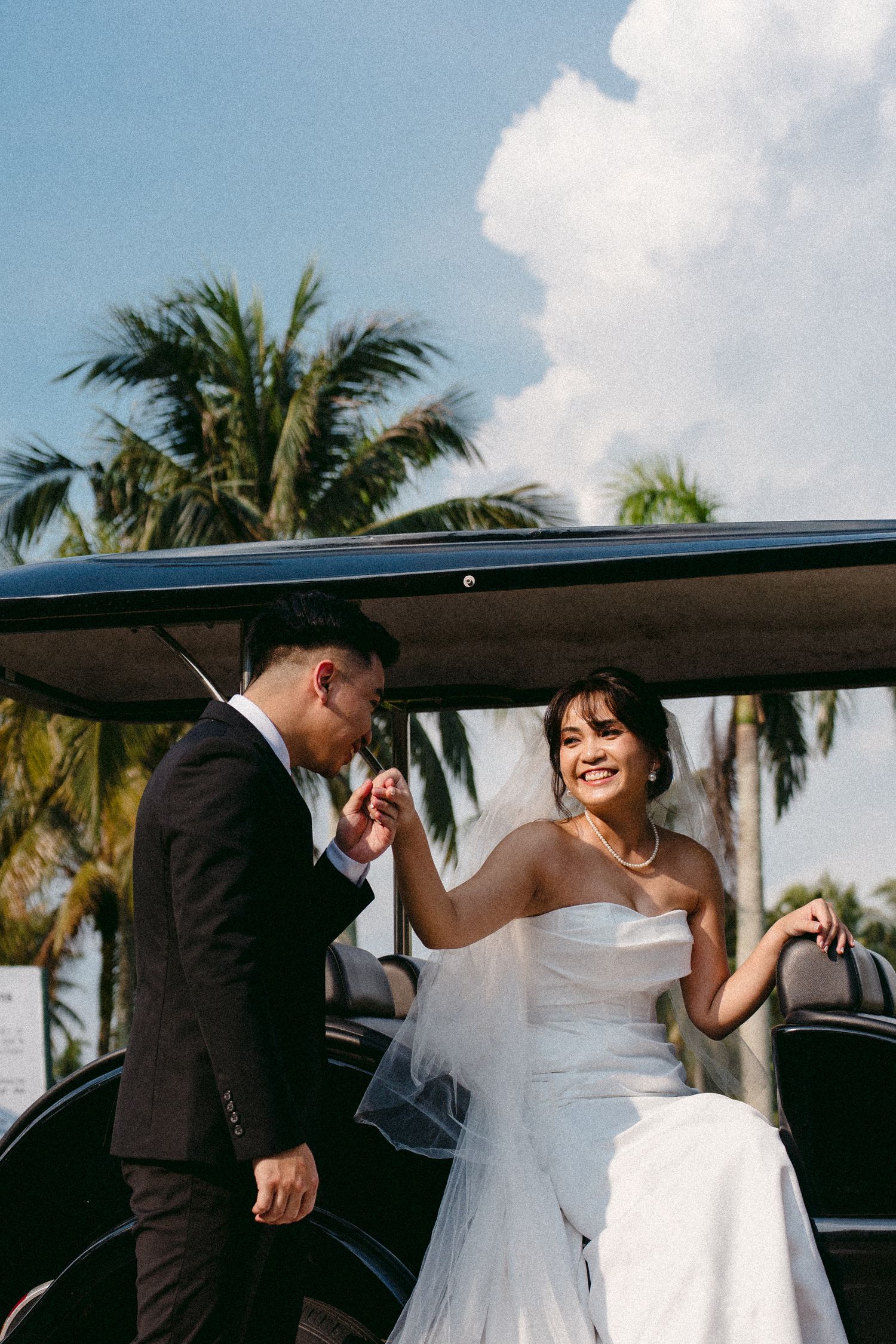 A romantic wedding moment between a bride in white dress and groom beside a vintage car with palm trees in the background.