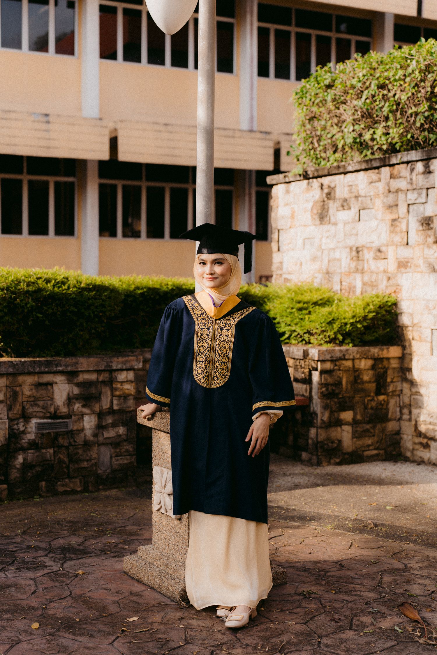 A graduate in traditional academic regalia with gold embroidered details stands on stone pavement near a building.
