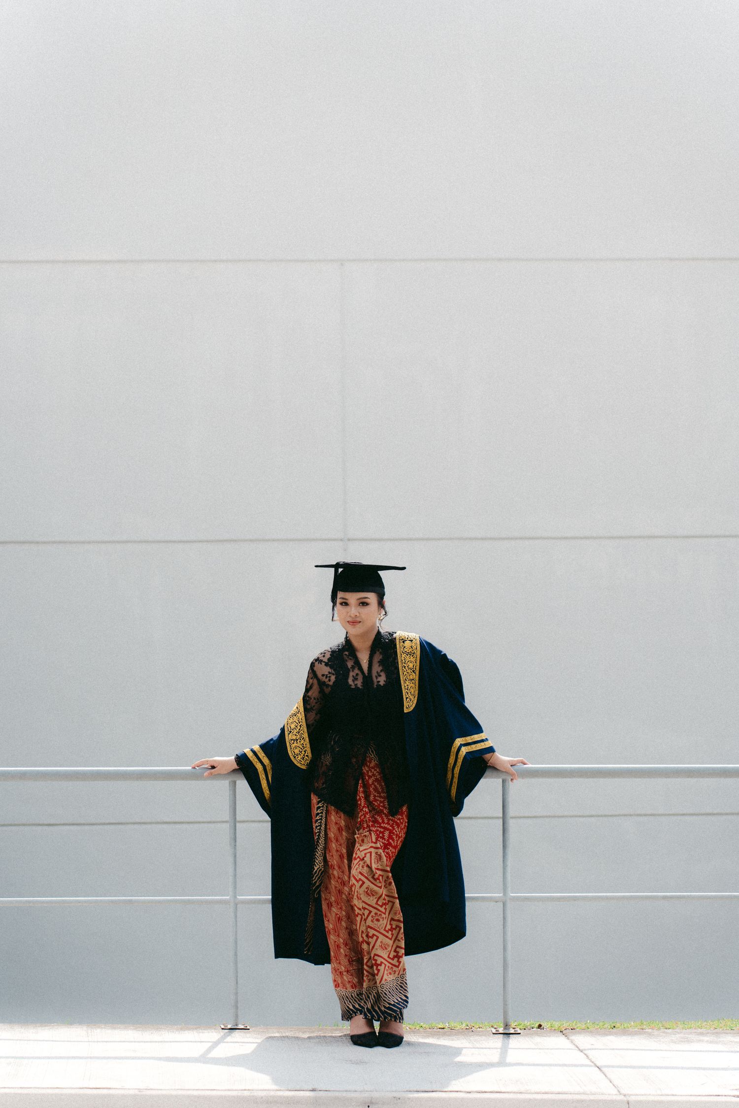 Graduate in black academic regalia with gold trim and patterned pants stands against a white wall.