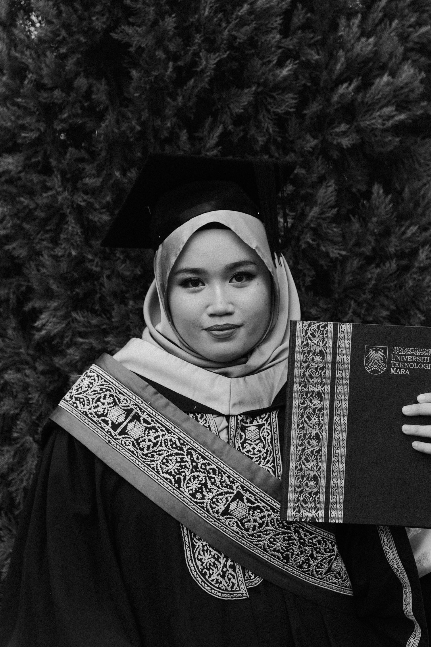 A graduate in traditional academic regalia poses with diploma against foliage background in black and white photography.