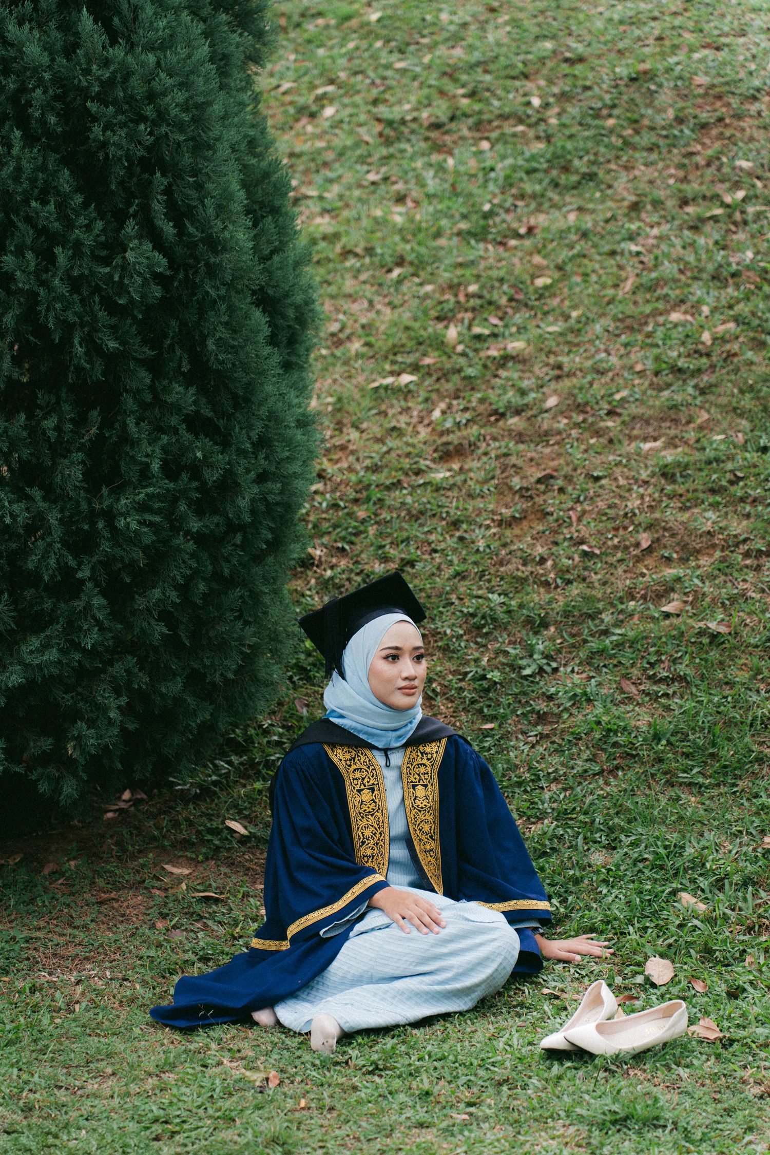 Person in traditional embroidered vest and navy robe sits cross-legged on grass near an evergreen tree with ballet shoes nearby.