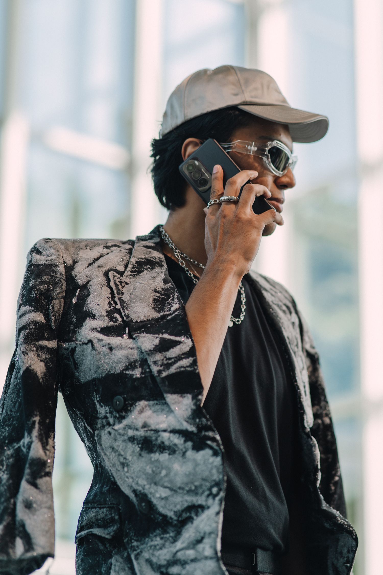 Stylish person in cap and sunglasses speaks on phone wearing abstract print jacket.