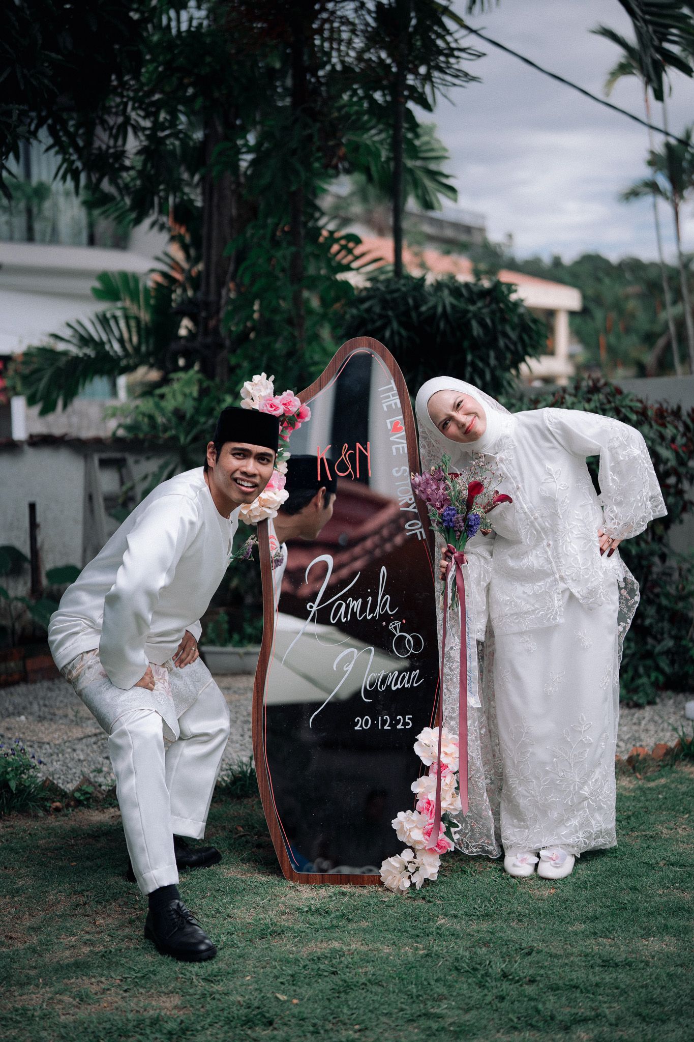 Muslim couple in white wedding attire posing beside an oval mirror sign displaying their names and date 20.12.25.