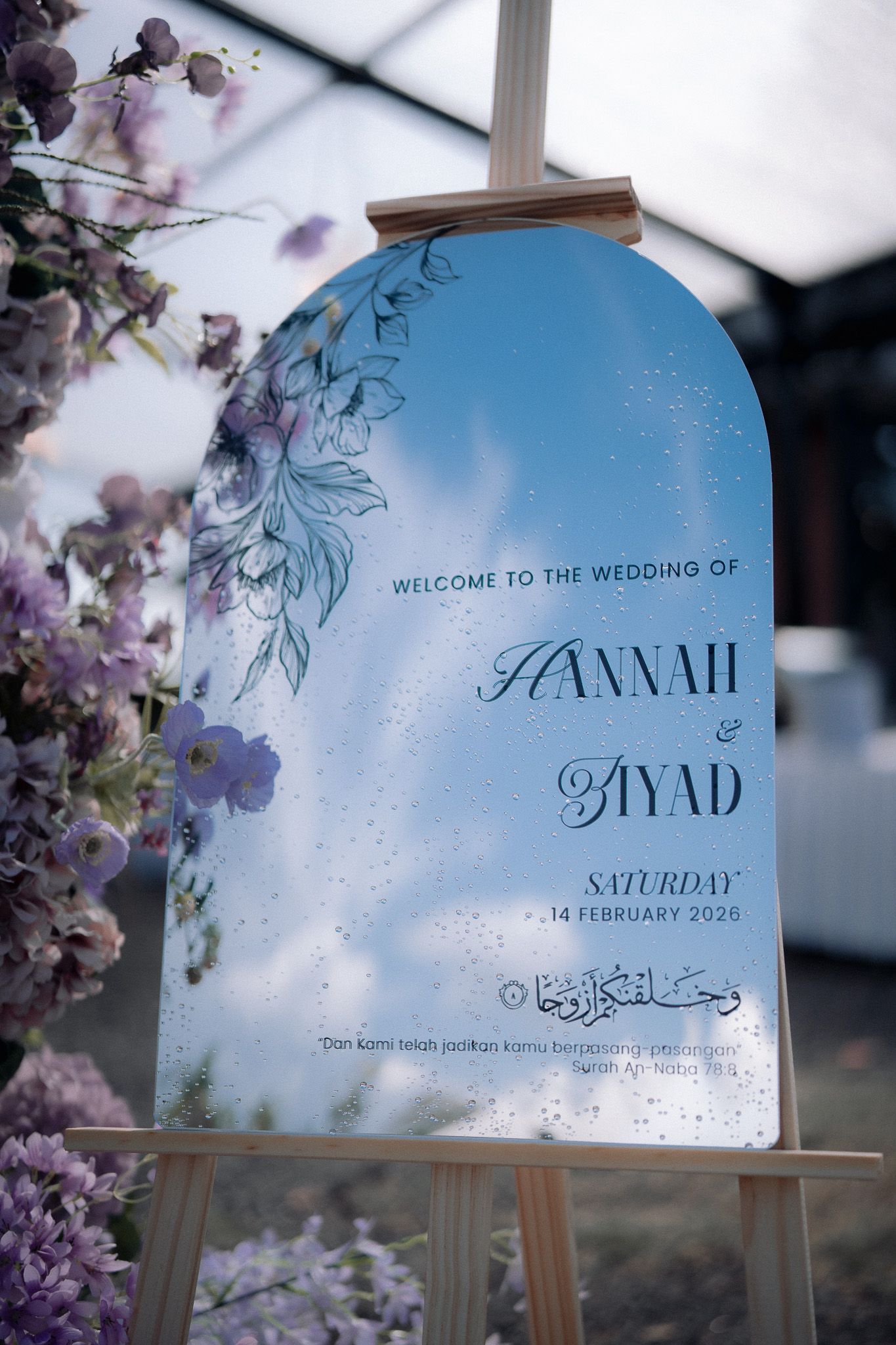Elegant wedding welcome sign for Hannah and Ayad on February 14, 2026, featuring floral decor and blue sky design.