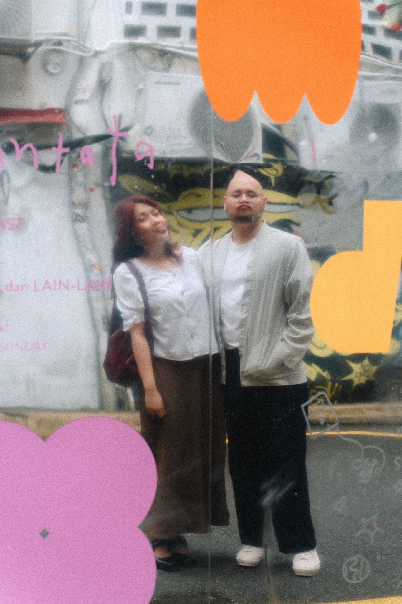 A man and woman pose together in front of a colorful mural wall with orange and pink flower decorations.