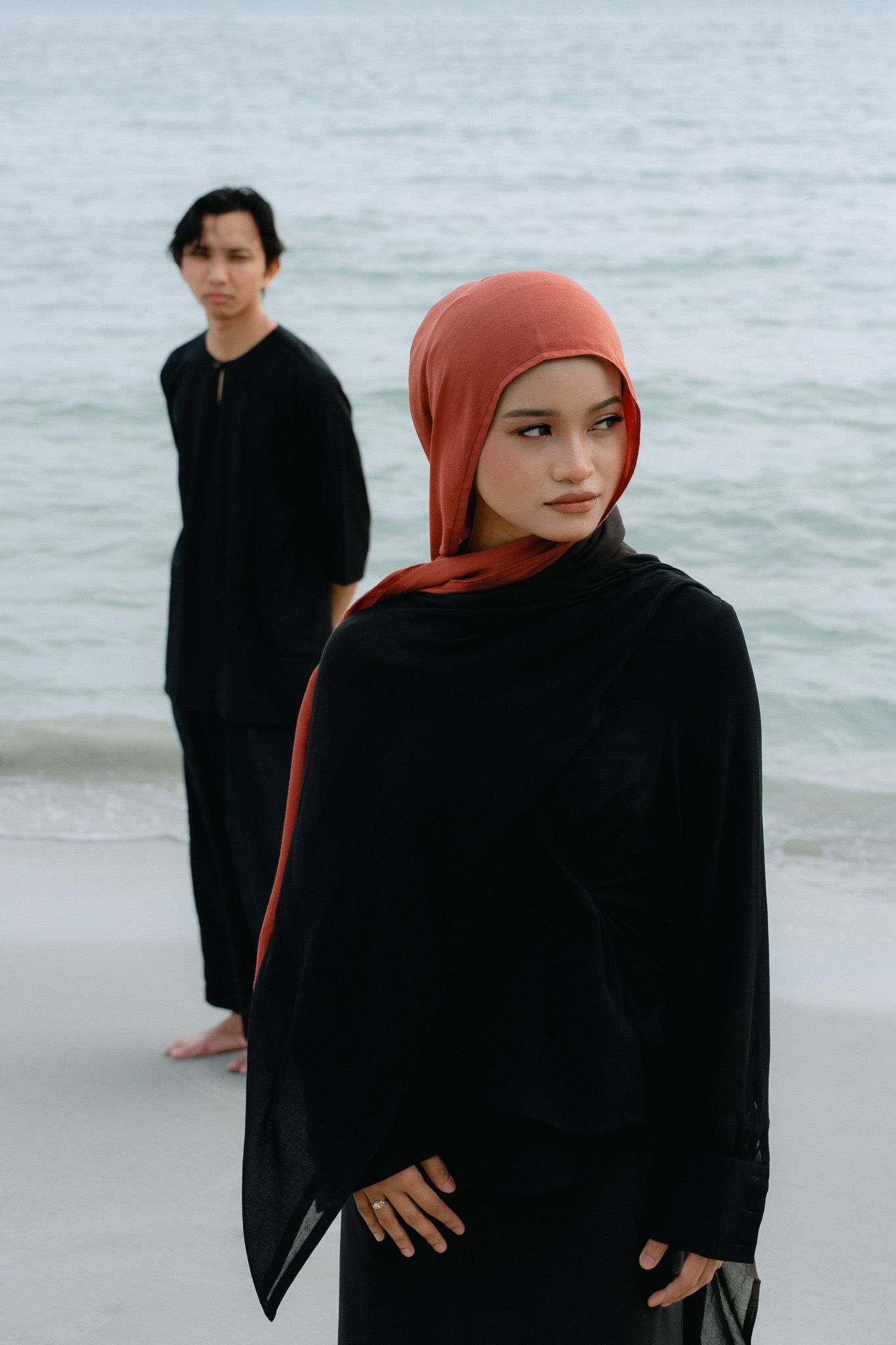 A woman in black abaya and rust hijab poses on a beach with a man in black standing behind her.