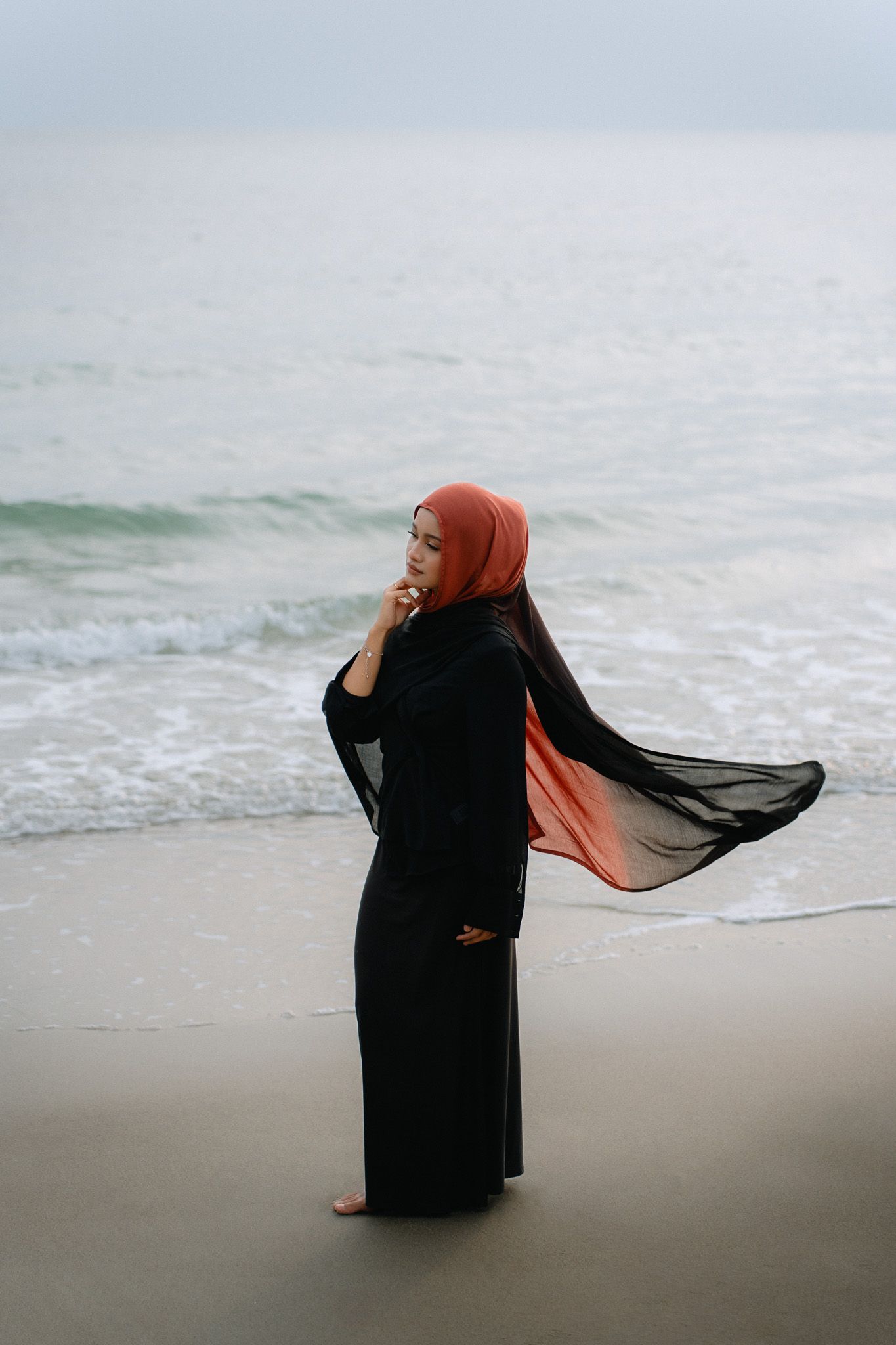 Woman in black abaya and orange hijab standing on beach with ocean waves in background.