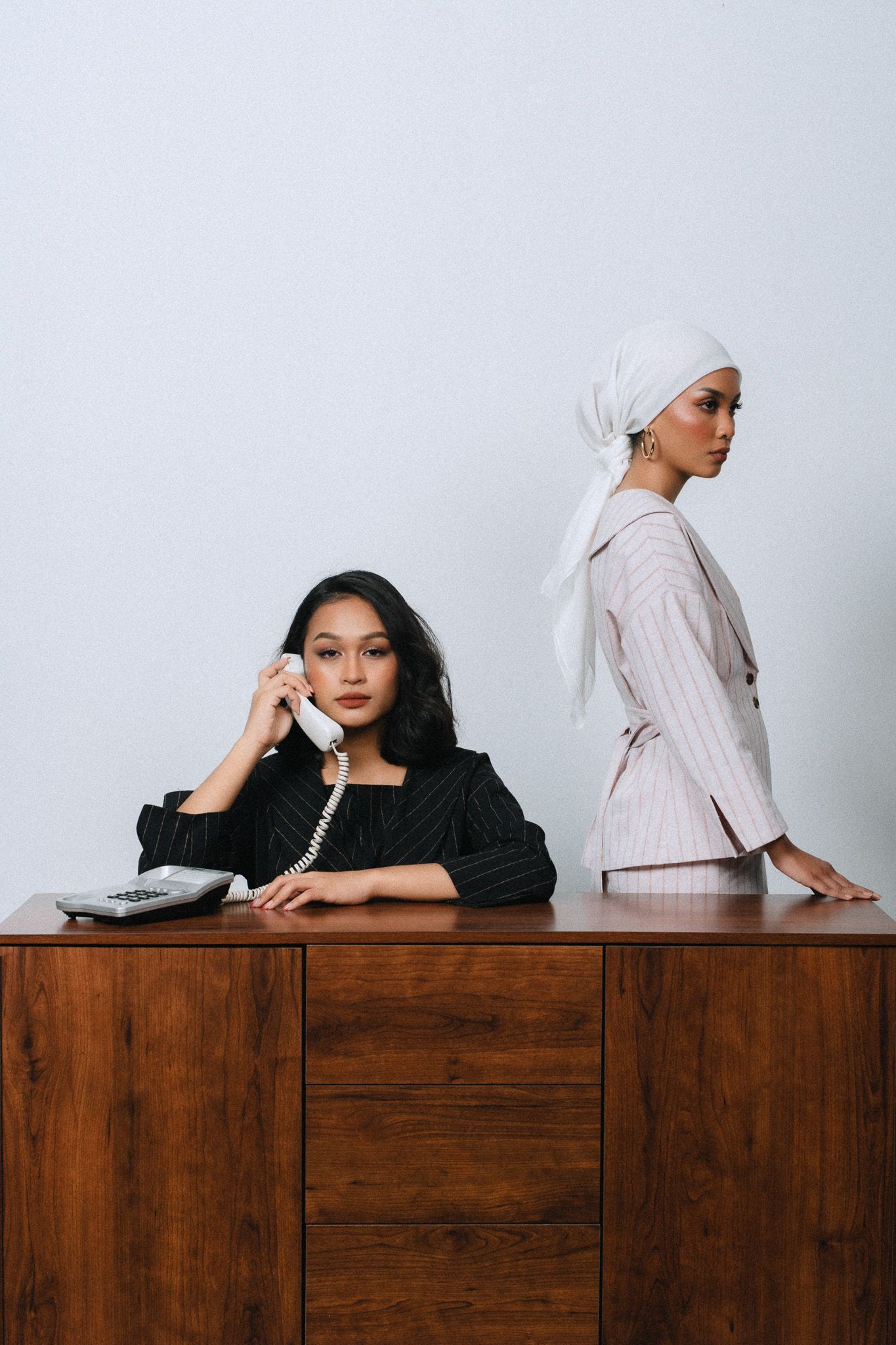 Two women pose near a wooden desk, one in black holding a phone, the other in a pink suit and white head wrap.