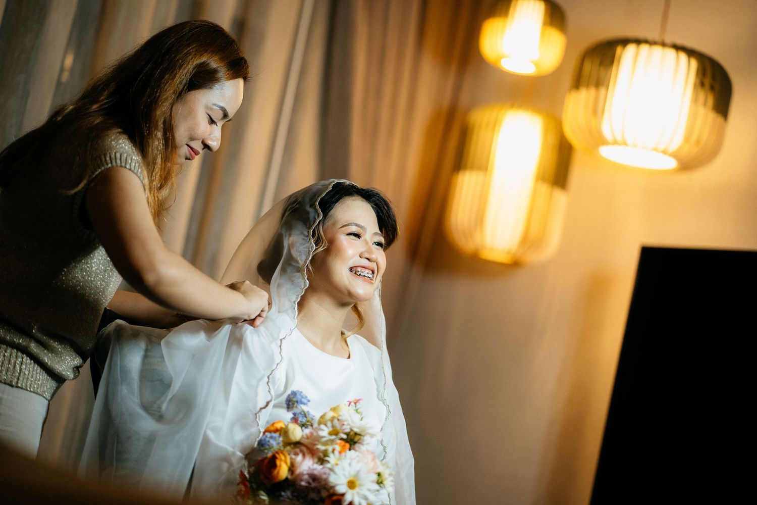 A person getting their hair styled while holding a floral bouquet in a warmly lit room with hanging pendant lamps.