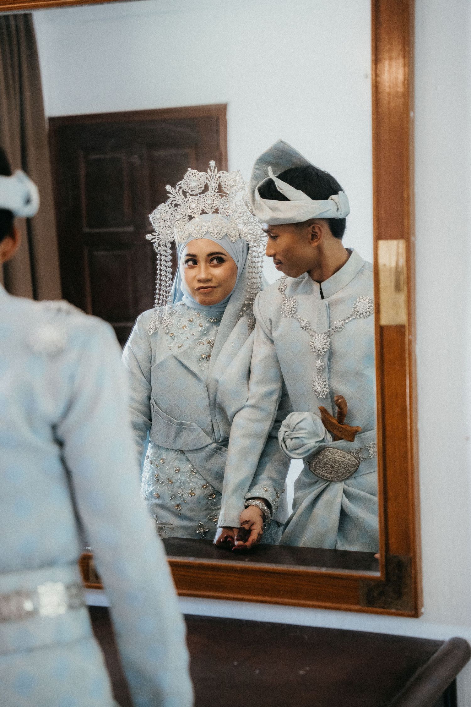 Traditional wedding couple in matching silver-gray attire reflected in mirror, with bride wearing ornate beaded headdress.