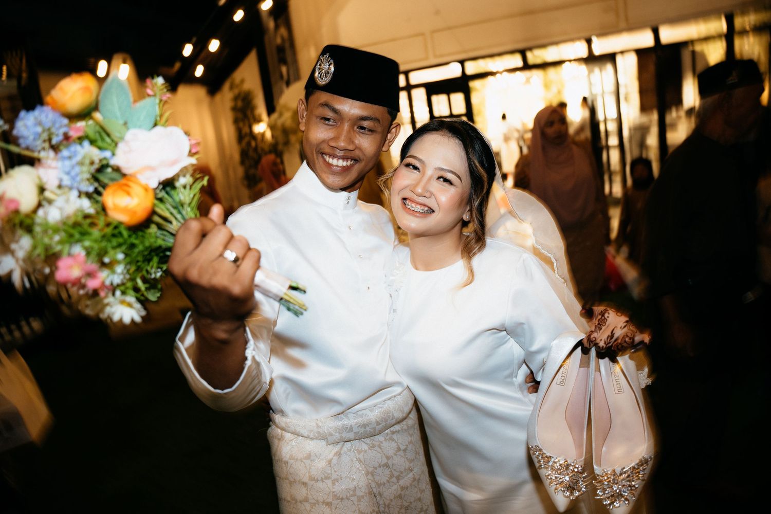 A happy couple in traditional wedding attire smiles and celebrates at their evening reception.
