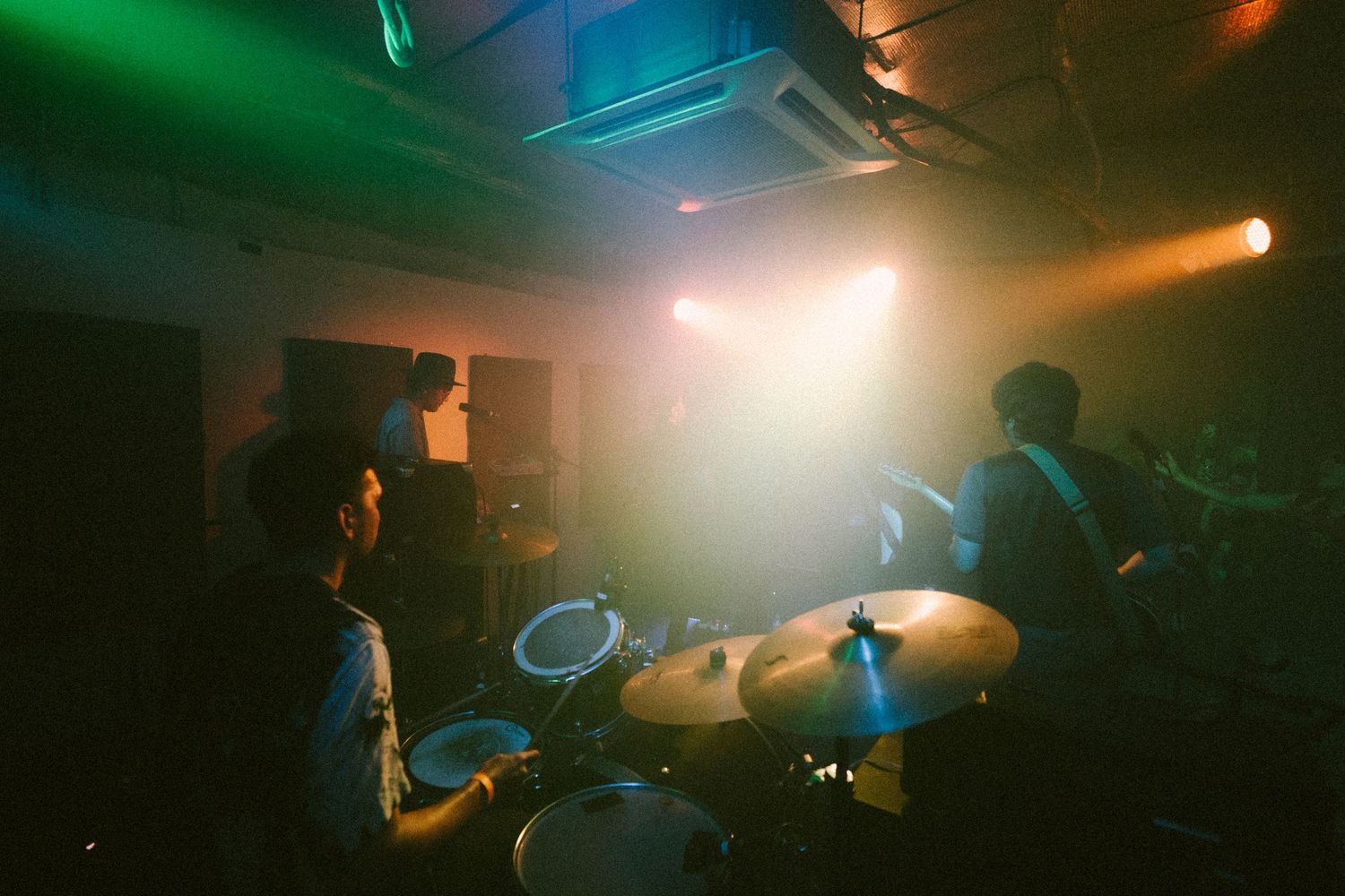 Musicians perform on stage in a dimly lit venue with green and orange atmospheric lighting during a live show.