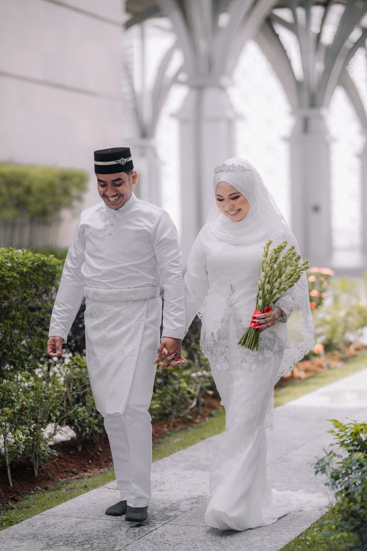 A newlywed couple in traditional white Malay wedding attire walking together in a garden with ornate archways.