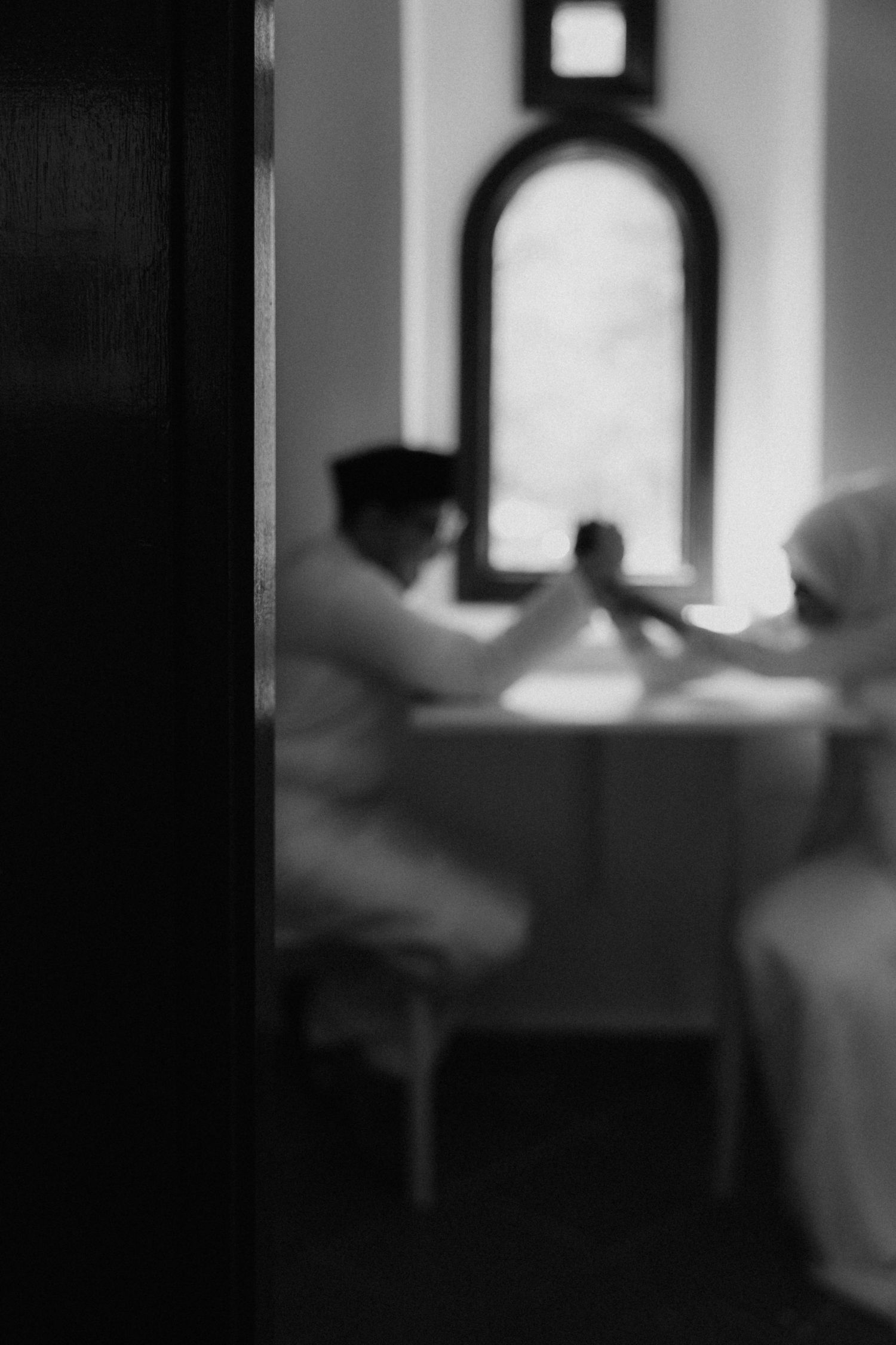 A blurry black and white architectural photograph showing figures seated at a table near an arched window.