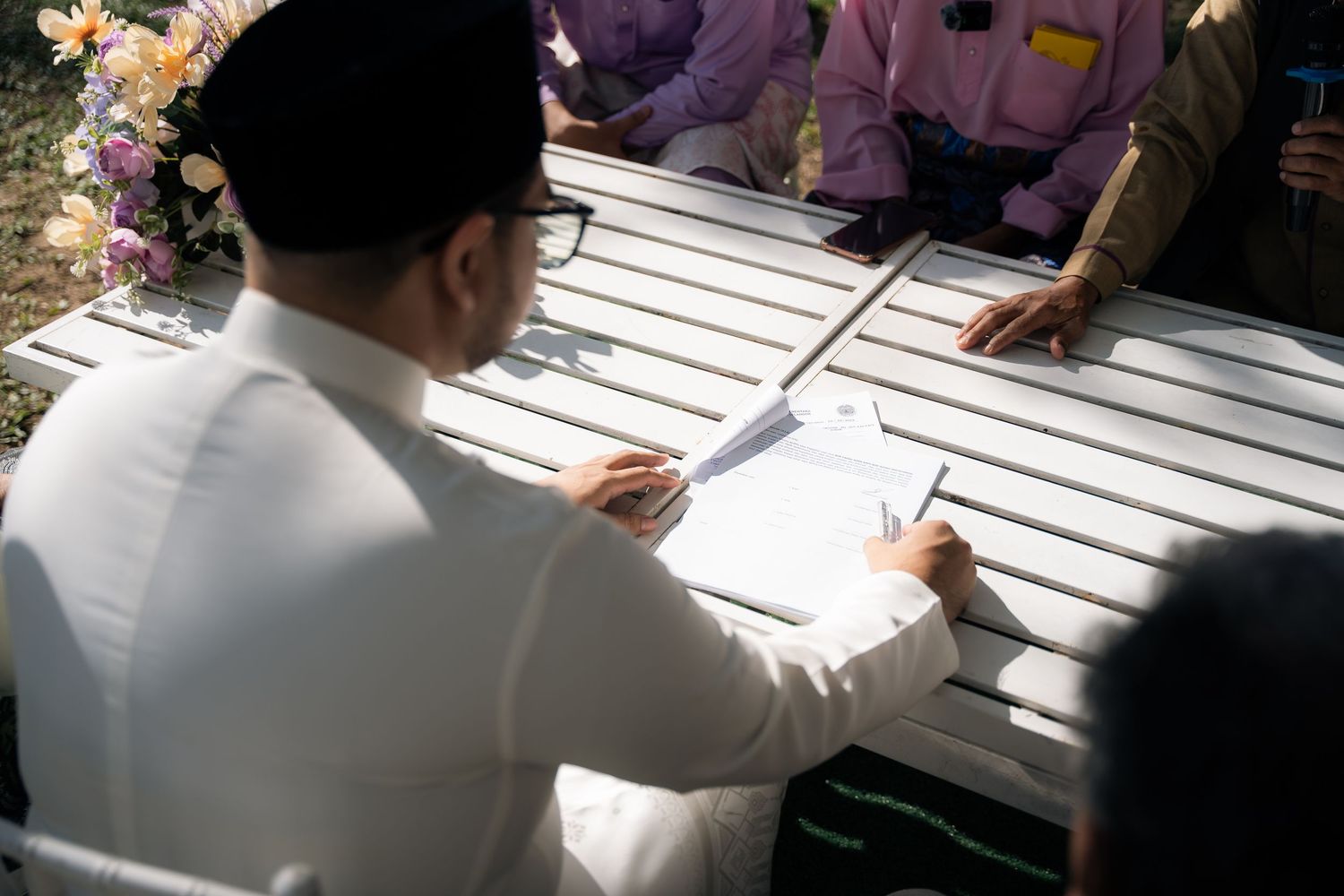 Person in white shirt signs document at outdoor table while others look on.