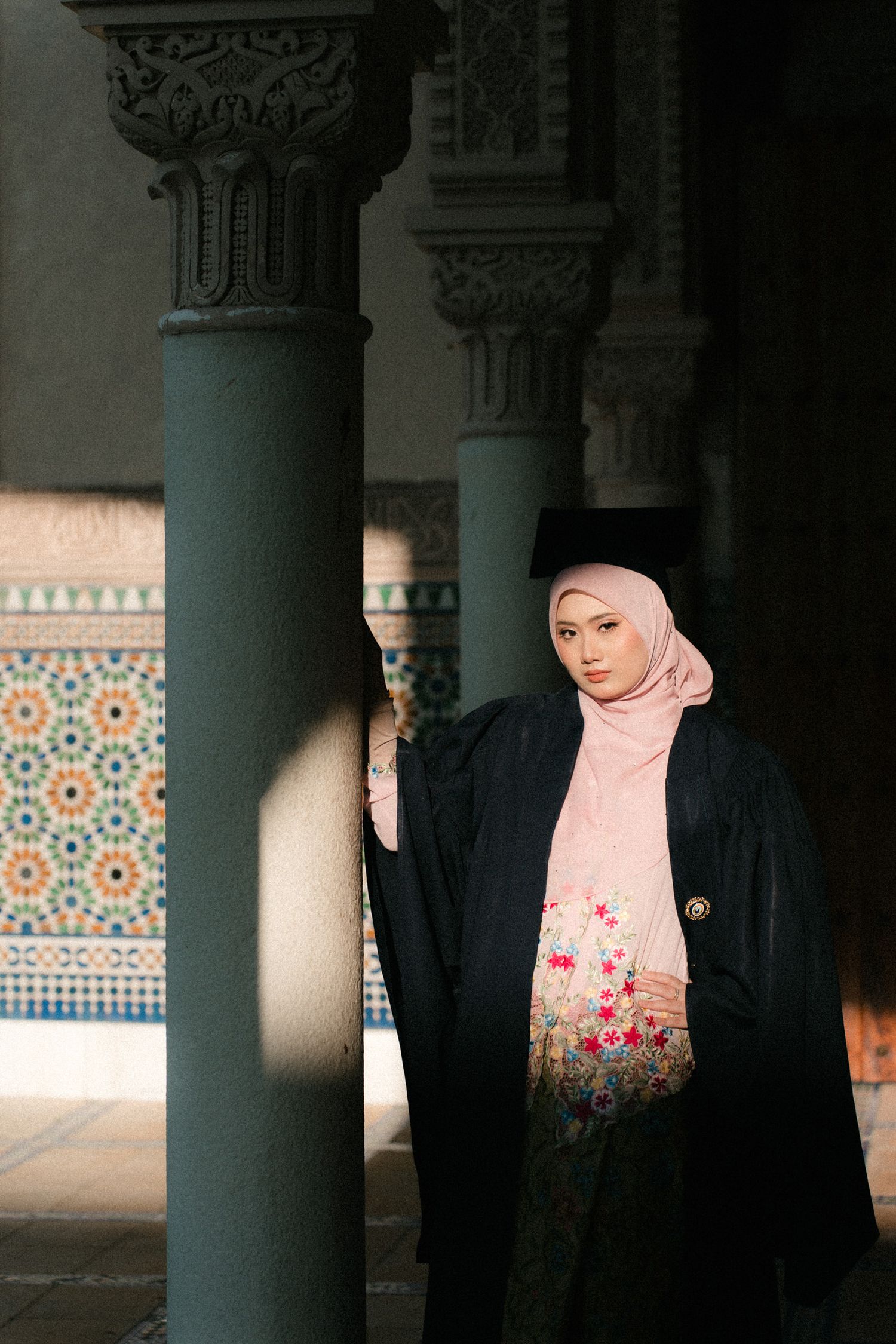 A person wearing a pink floral hijab stands between ornate columns with decorative Islamic architectural details in the background.