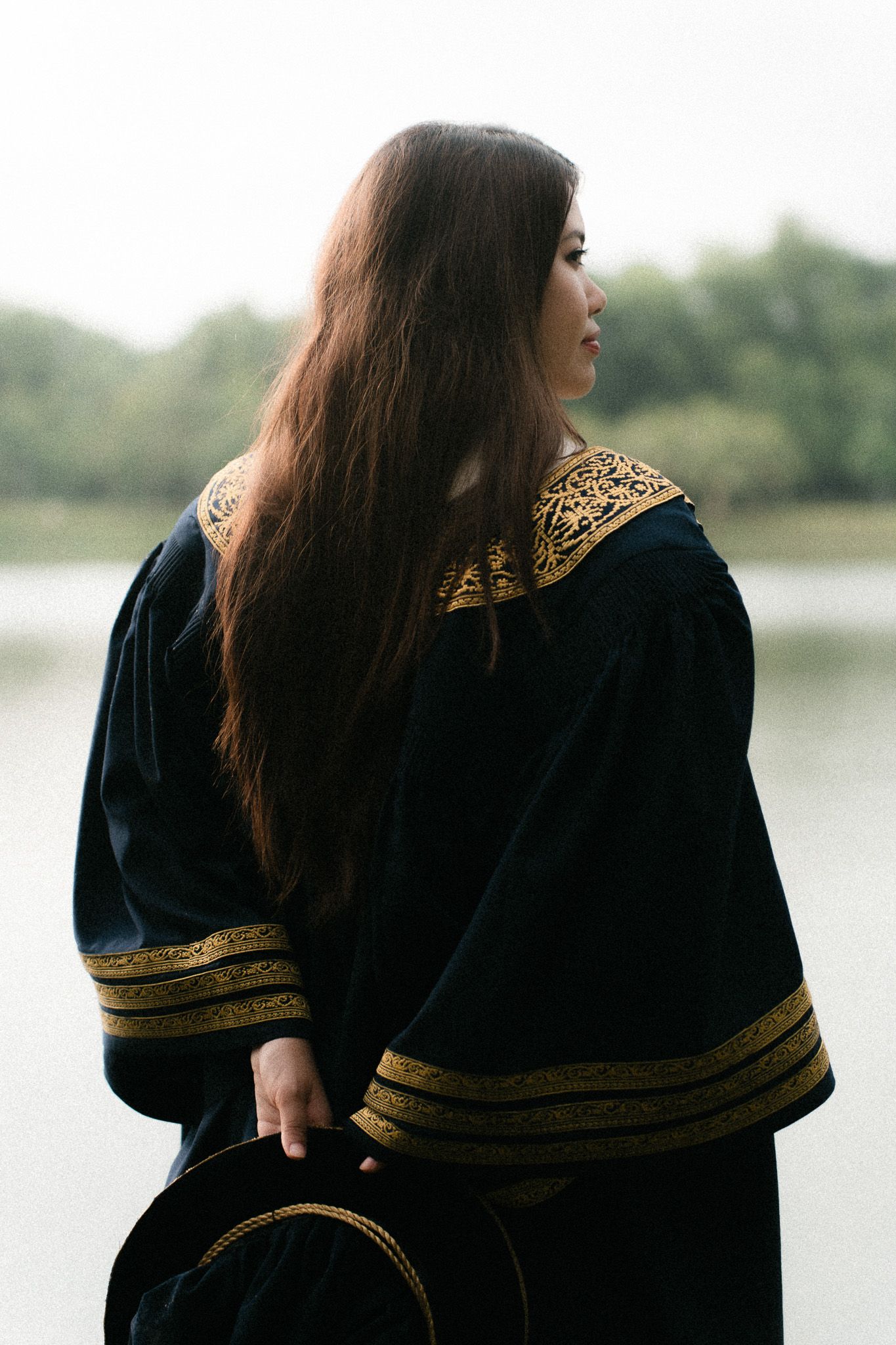 Woman in black and gold embroidered robe with long brown hair holds hat near misty lake.