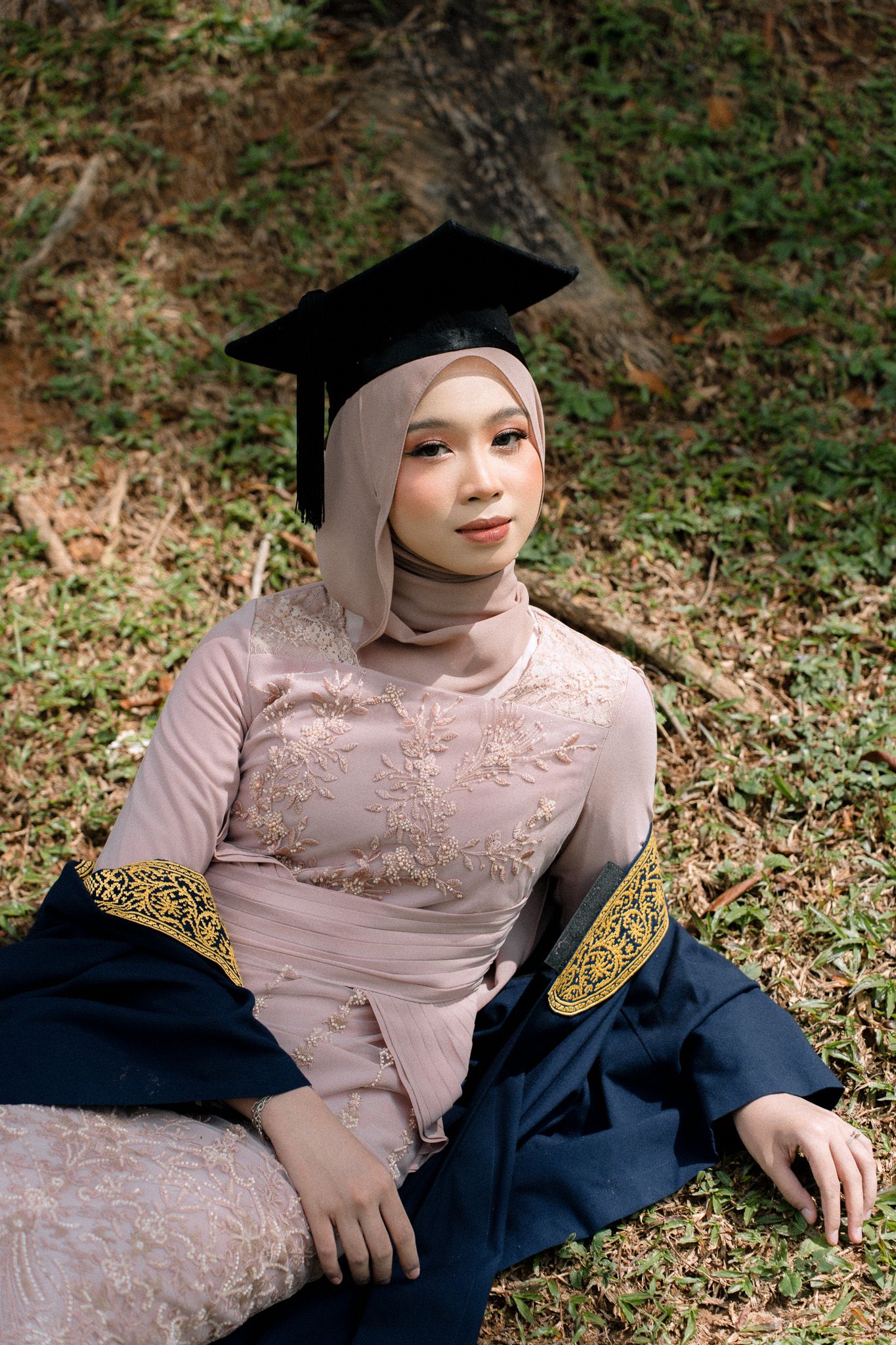 Muslim woman in hijab and graduation cap sits on grass in elegant embroidered kebaya, celebrating academic achievement outdoors.