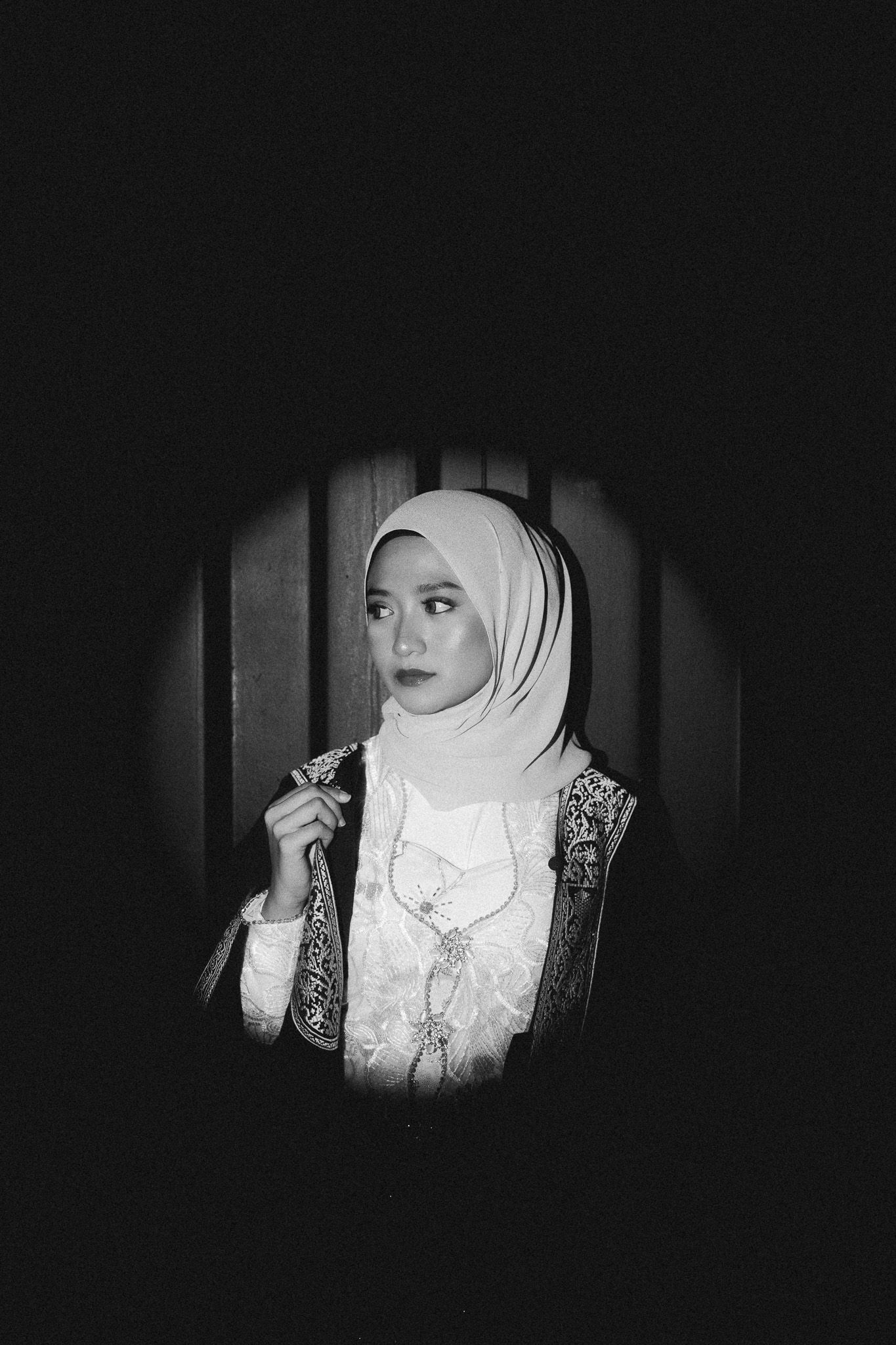 Black and white portrait of a woman wearing a hijab and traditional embroidered attire, posed in a dark, dramatic setting.