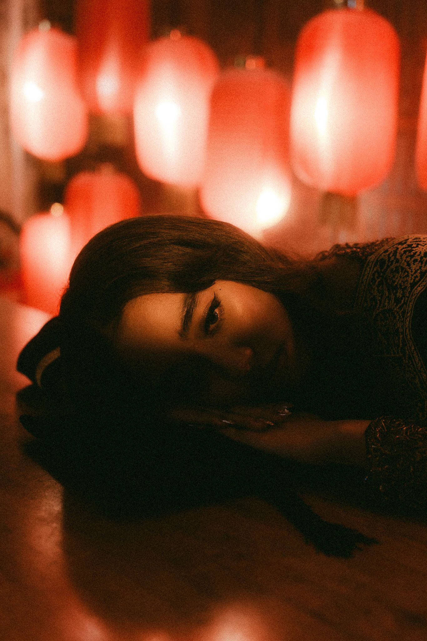 Woman lying on floor bathed in red glow from hanging Chinese lanterns in a moody, atmospheric portrait.