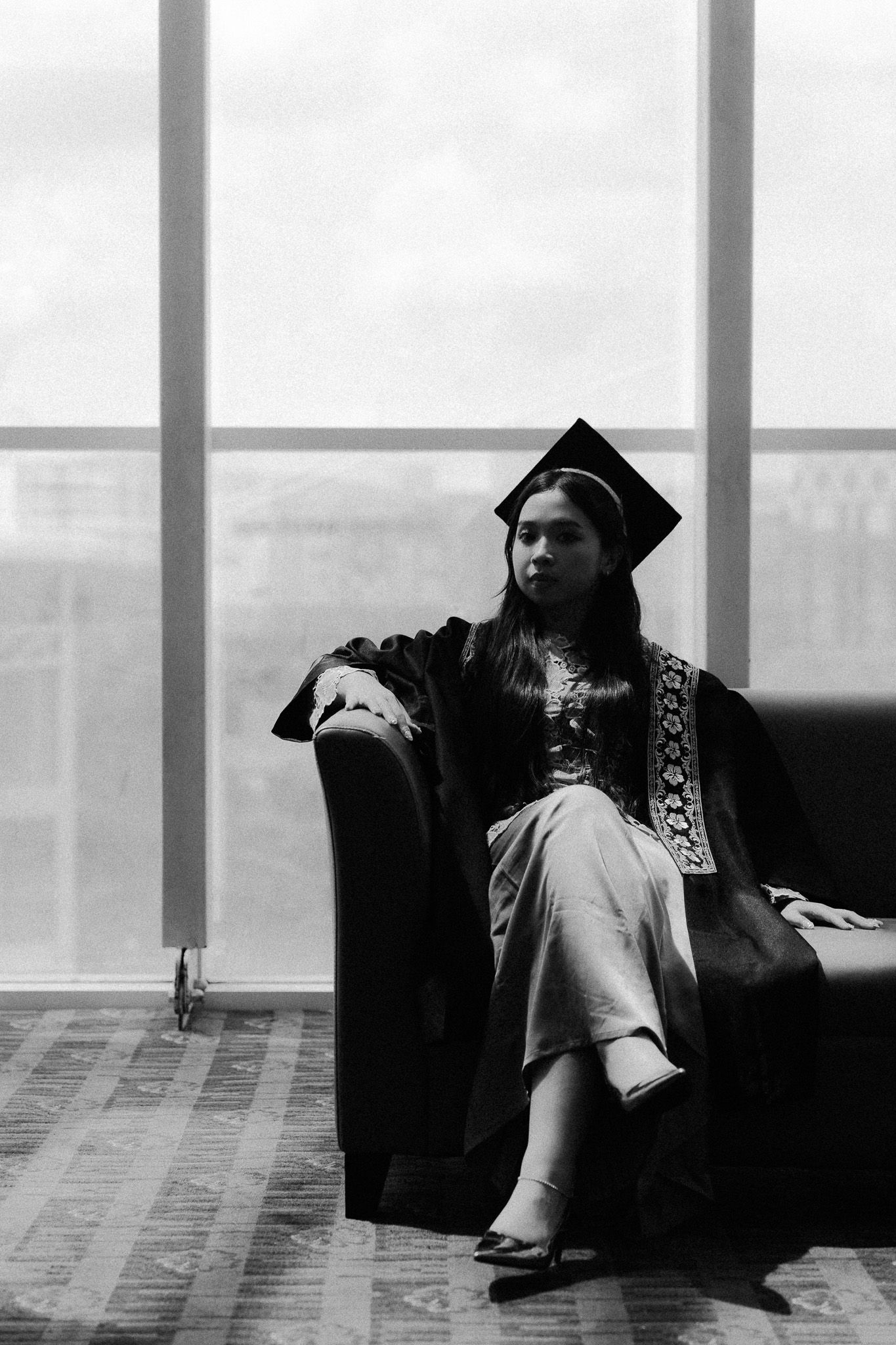 Graduate in cap and gown sitting confidently in a modern building with large windows, black and white portrait.
