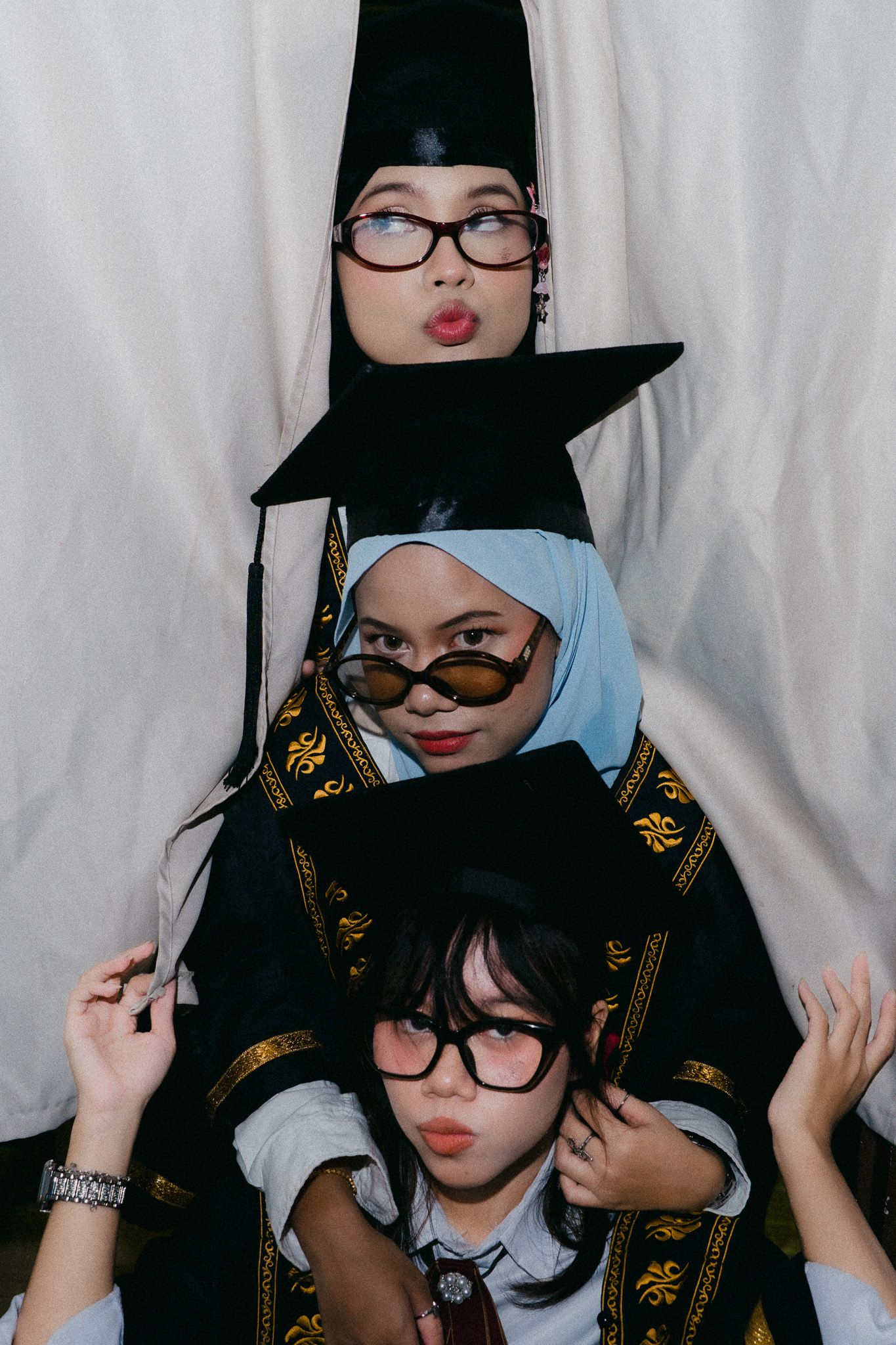 Three women in graduation caps and gowns with glasses making funny faces, stacked portrait pose.