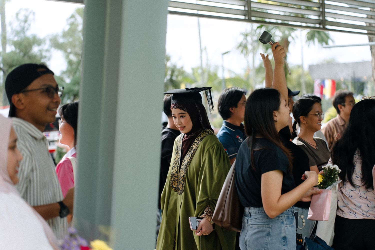 Graduate in cap and gown stands among crowd at outdoor graduation ceremony celebration.