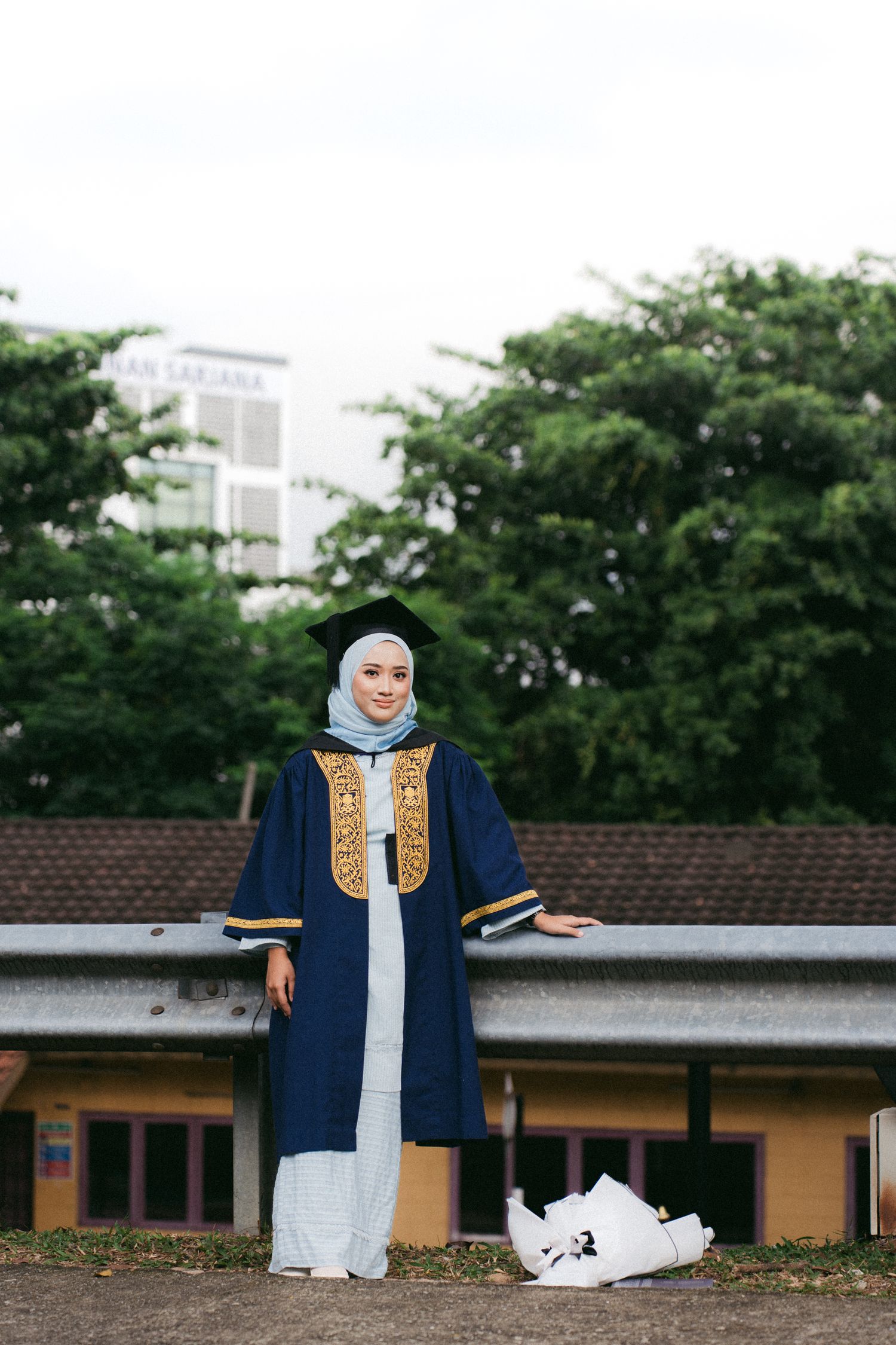 Person wearing navy blue graduation gown with gold accents and light hijab poses outside near trees and buildings.