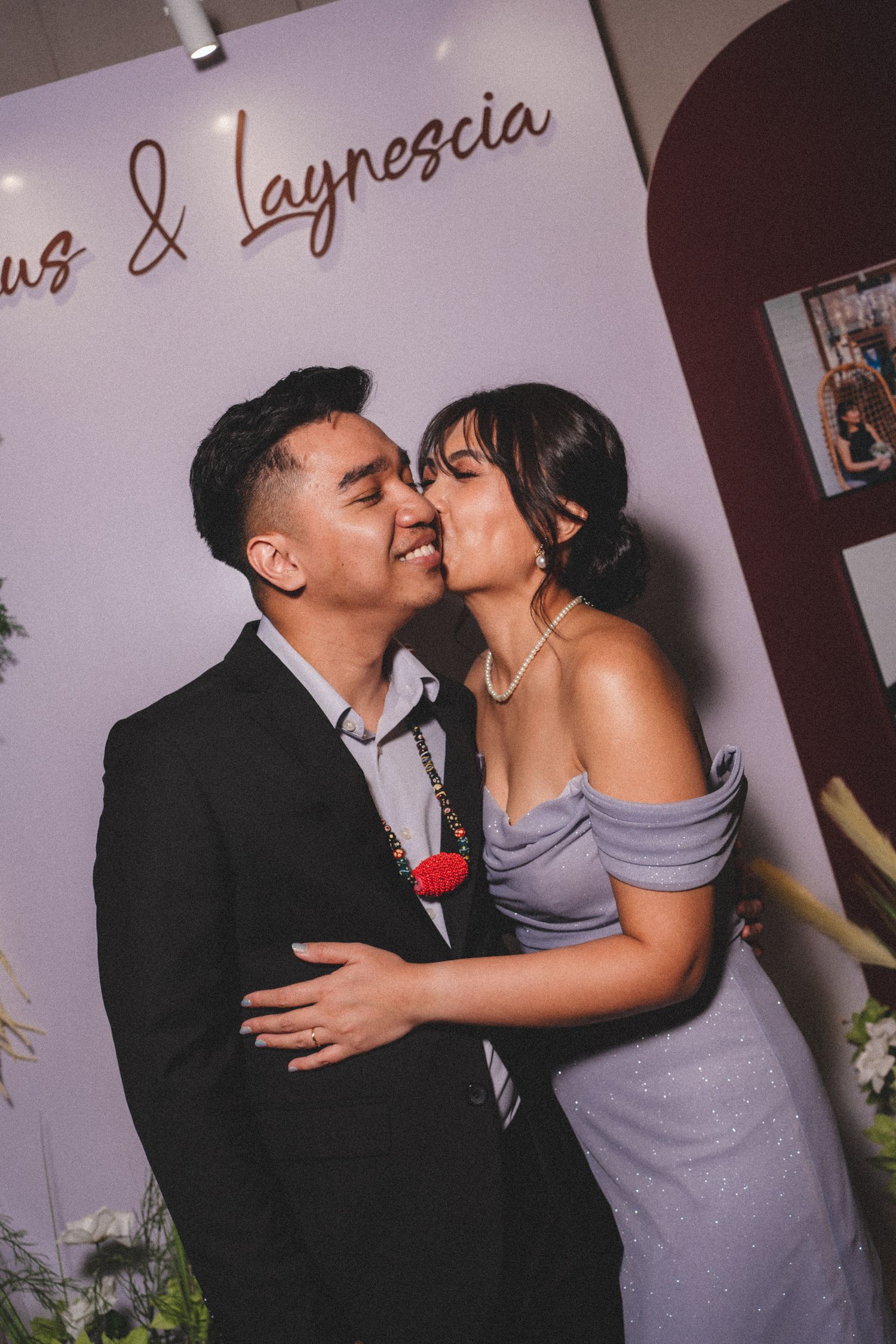 A romantic couple embraces and kisses in front of a decorated wedding photo backdrop with script text.