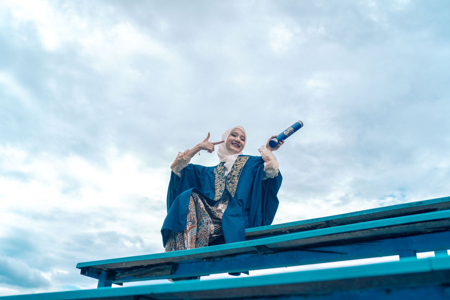 Graduate in blue academic regalia stands triumphantly on elevated platform against cloudy sky.
